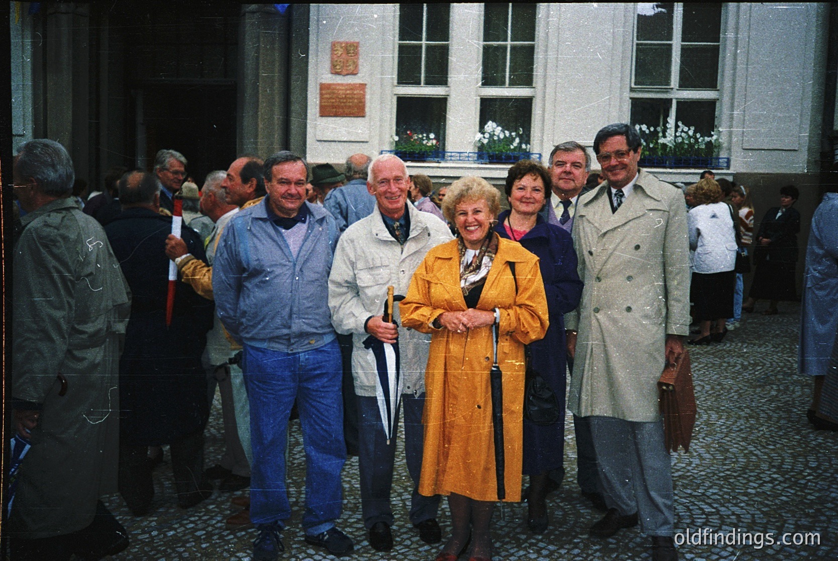 Group portrait in front of a neoclassical building with cobblestone courtyard, likely 1970s–1980s. Six adults pose in vintage outerwear—jackets, coats, and a cane—suggesting a formal or celebratory event. Crowd and architectural details hint at European setting.