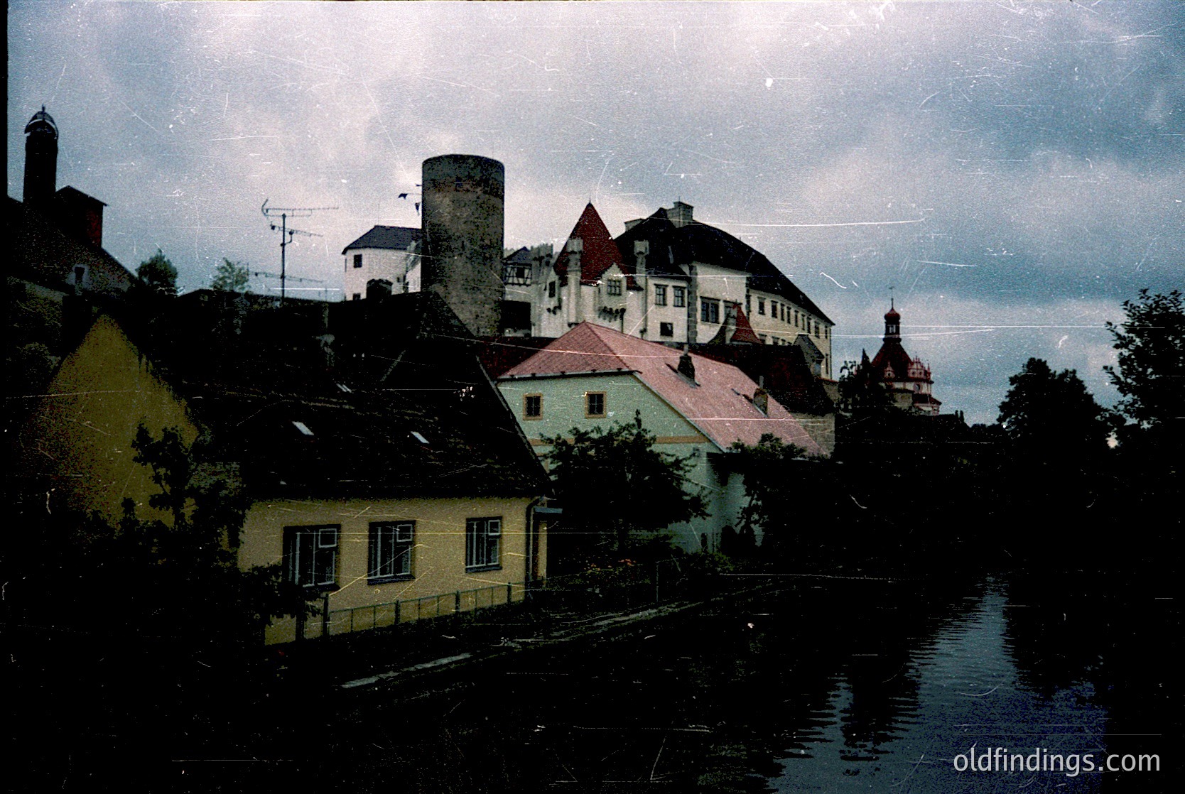 Vintage sepia-toned riverside town with historic European architecture. Prominent brick chimney and red-roofed buildings frame a canal, likely 20th-century. Overcast sky enhances nostalgic atmosphere.