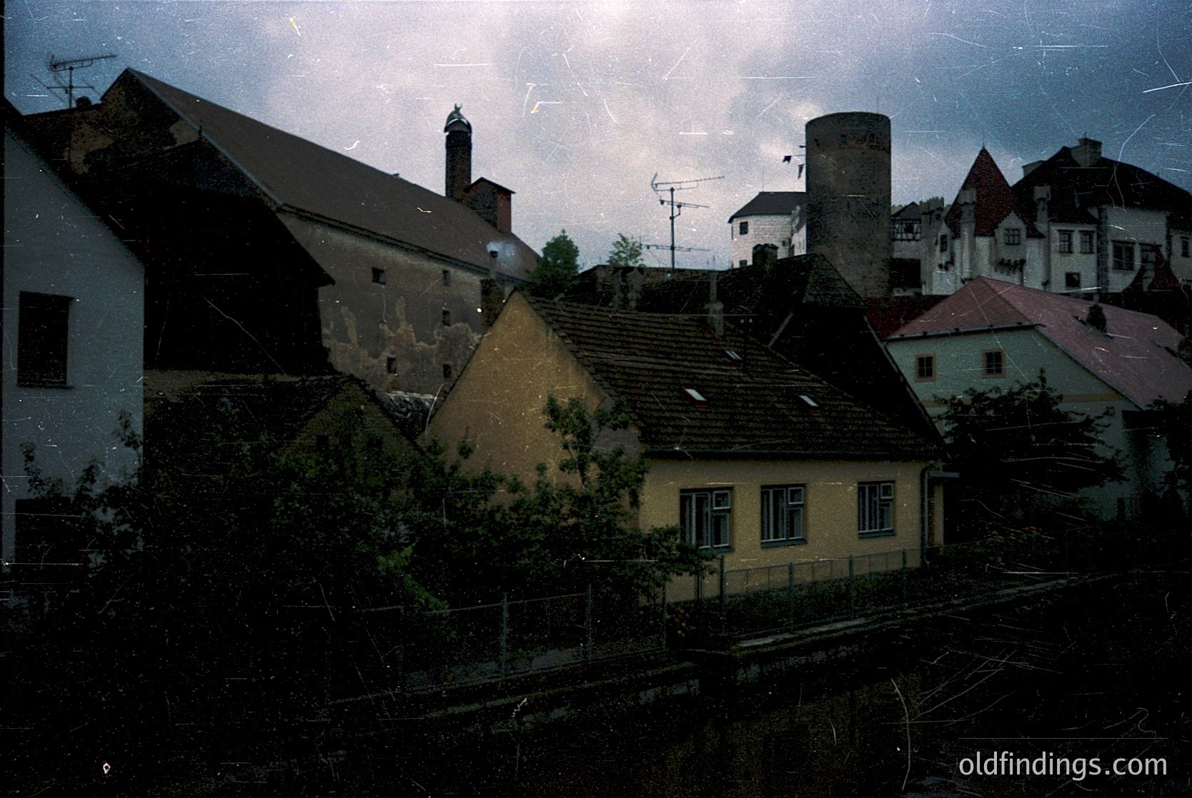 Vintage urban rooftops with aged, sloped shingles and chimneys under stormy skies. Mid-20th century European architecture—likely —shows weathered facades and industrial chimney. Overgrown greenery and rusted railings hint at neglected infrastructure.