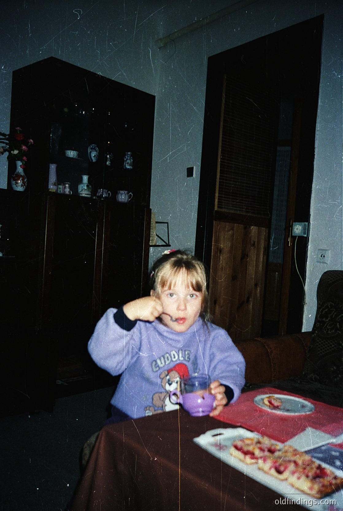 Young child in a 1990s-style "Cuddle" hoodie poses at a table with a small cup, likely for a drink or snack. Background features a wooden cabinet with ceramic teacups, a vintage-style sofa, and a doorway. Warm, lived-in interior suggests a mid-20th-century home.