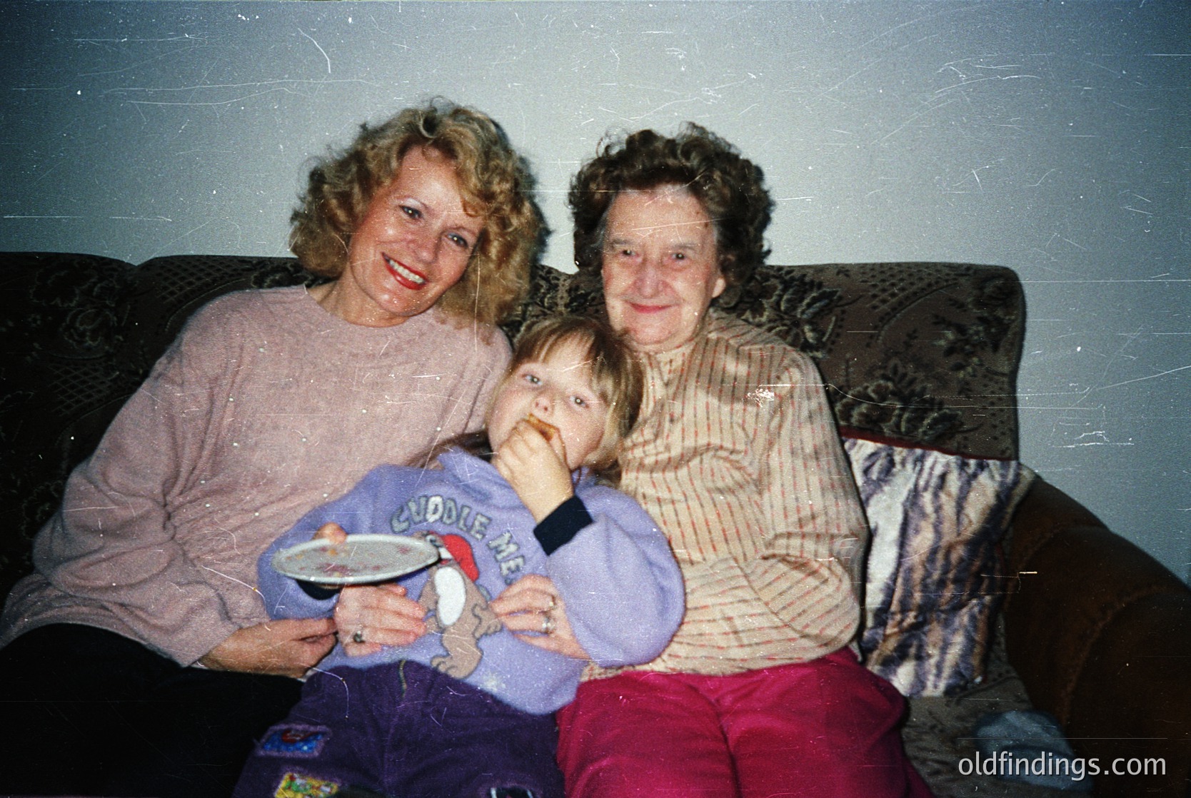 Vintage indoor portrait of three generations: two women (likely grandmother and mother) and a toddler in a "Cuddle Me" onesie. Warm, lived-in setting with patterned sofa and faded wallpaper. Mid-1990s American family life captured.