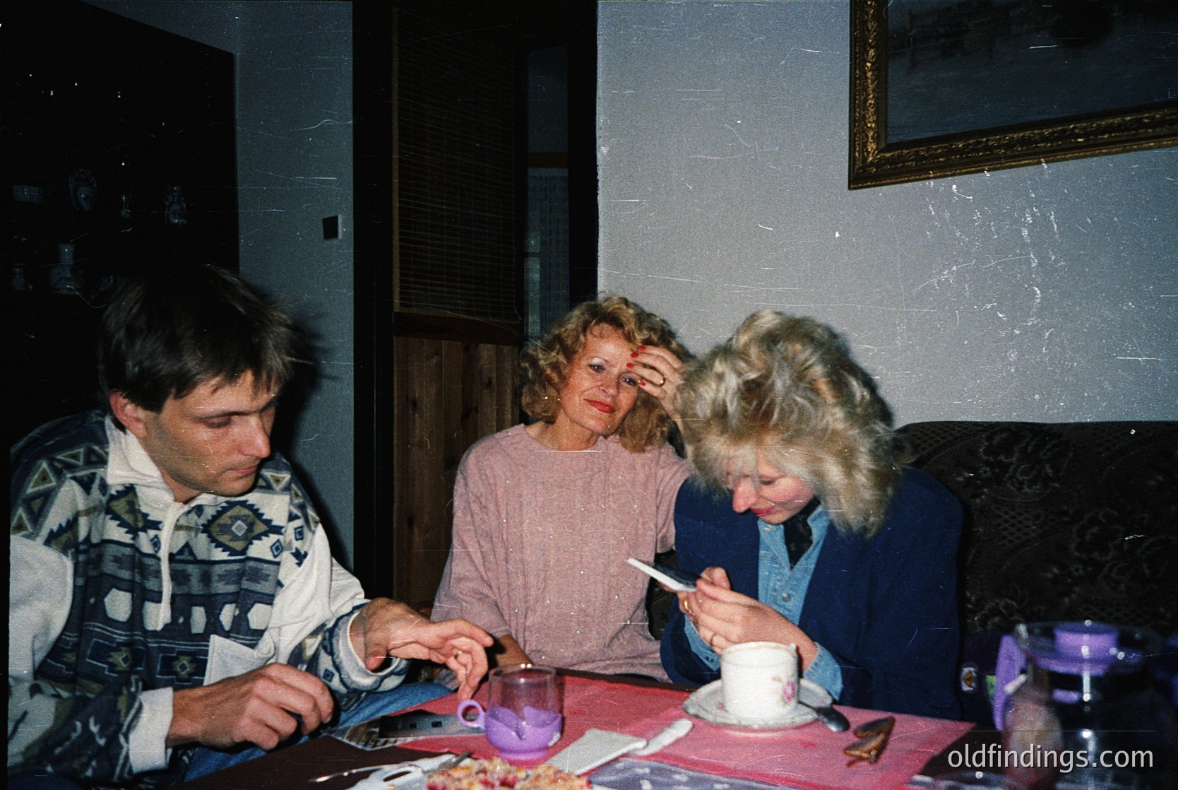 Three individuals seated indoors, likely mid-1990s, in a cozy, textured-wall setting. Left: man in patterned sweater, holding a magazine. Center: woman in pink sweater, resting hand on head. Right: woman in blue jacket, holding a cup. Table features a teapot, saucer, and scattered objects. Warm, intimate atmosphere.