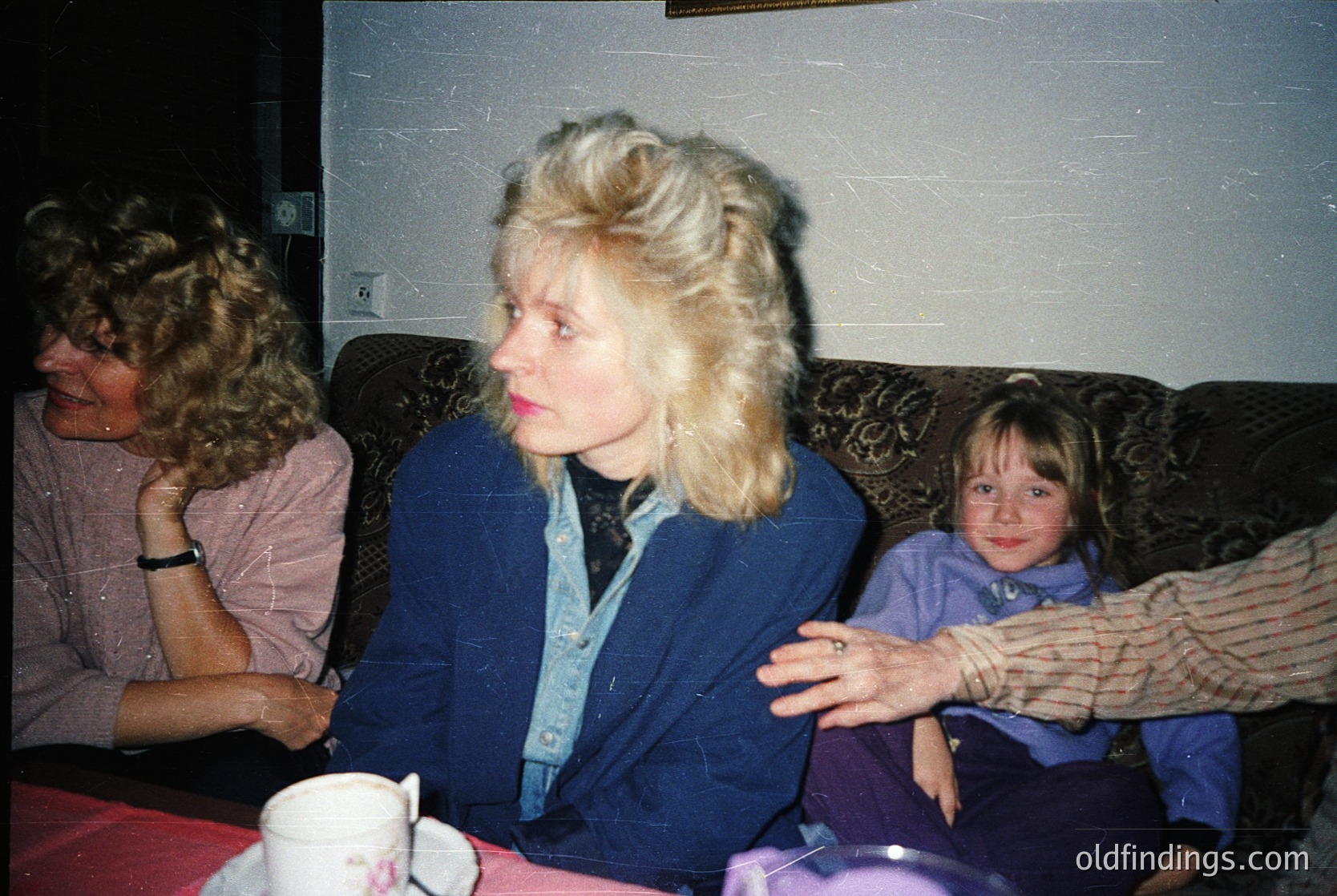 Vintage indoor portrait of three women in a dimly lit room, likely 1970s–1980s. The woman in the center wears a voluminous blue jacket and a checkered blouse, while the child on the right has a cropped haircut and a striped sweater. A teacup and saucer sit on a patterned tablecloth. Warm, nostalgic family moment.