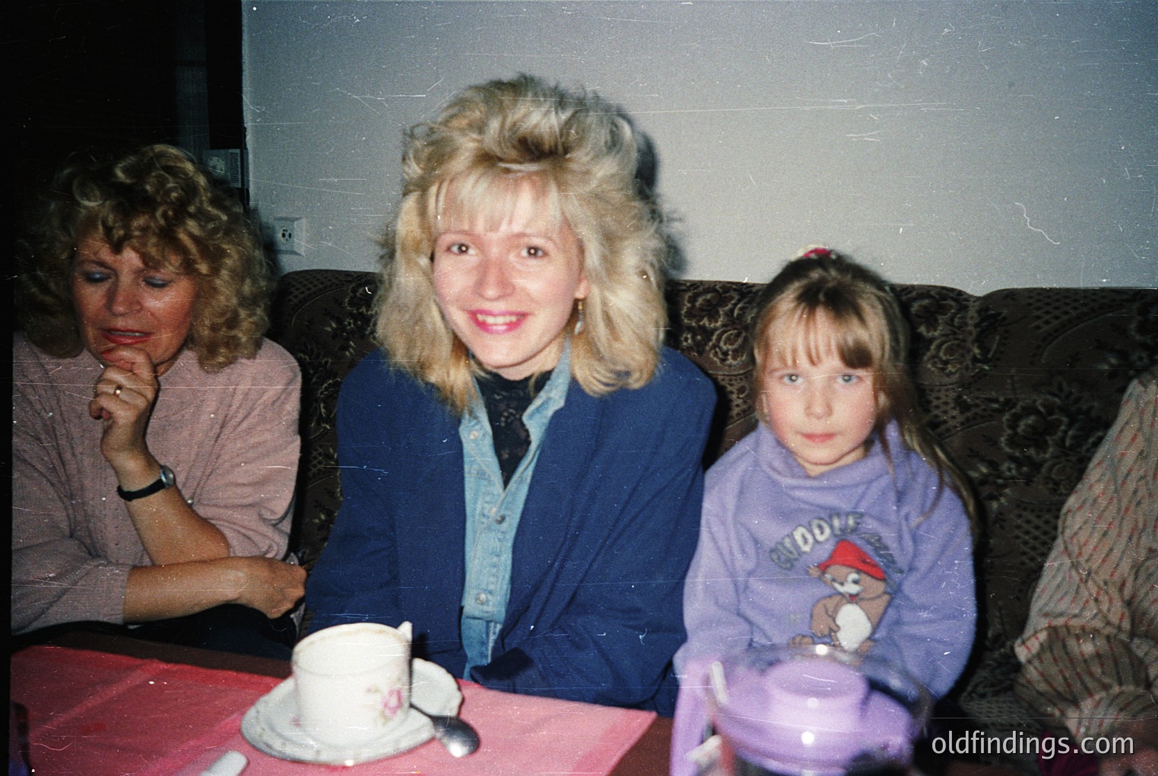 Three women pose indoors, likely 1980s–1990s. Center: woman in a blue cardigan with voluminous hair, smiling. Left: seated woman in a pink blouse, resting chin on hand. Right: child in a purple sweatshirt with a cartoon character, holding a cup. Vintage tableware and patterned upholstery suggest a casual, domestic setting.