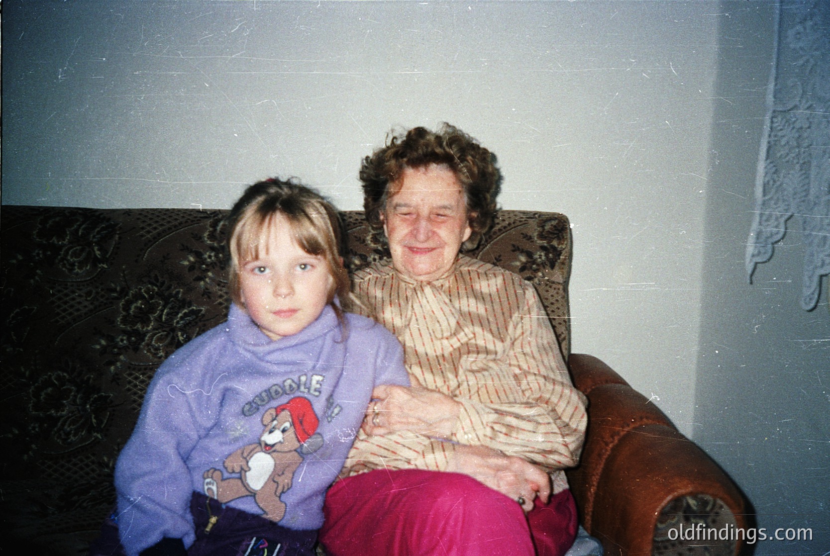 Vintage indoor portrait of a grandmother and young girl on a patterned armchair, likely 1980s–1990s. The girl wears a purple sweatshirt with a cartoon character and stuffed toy. The elder woman smiles warmly, holding the child close. Plain interior with floral wallpaper and simple decor.