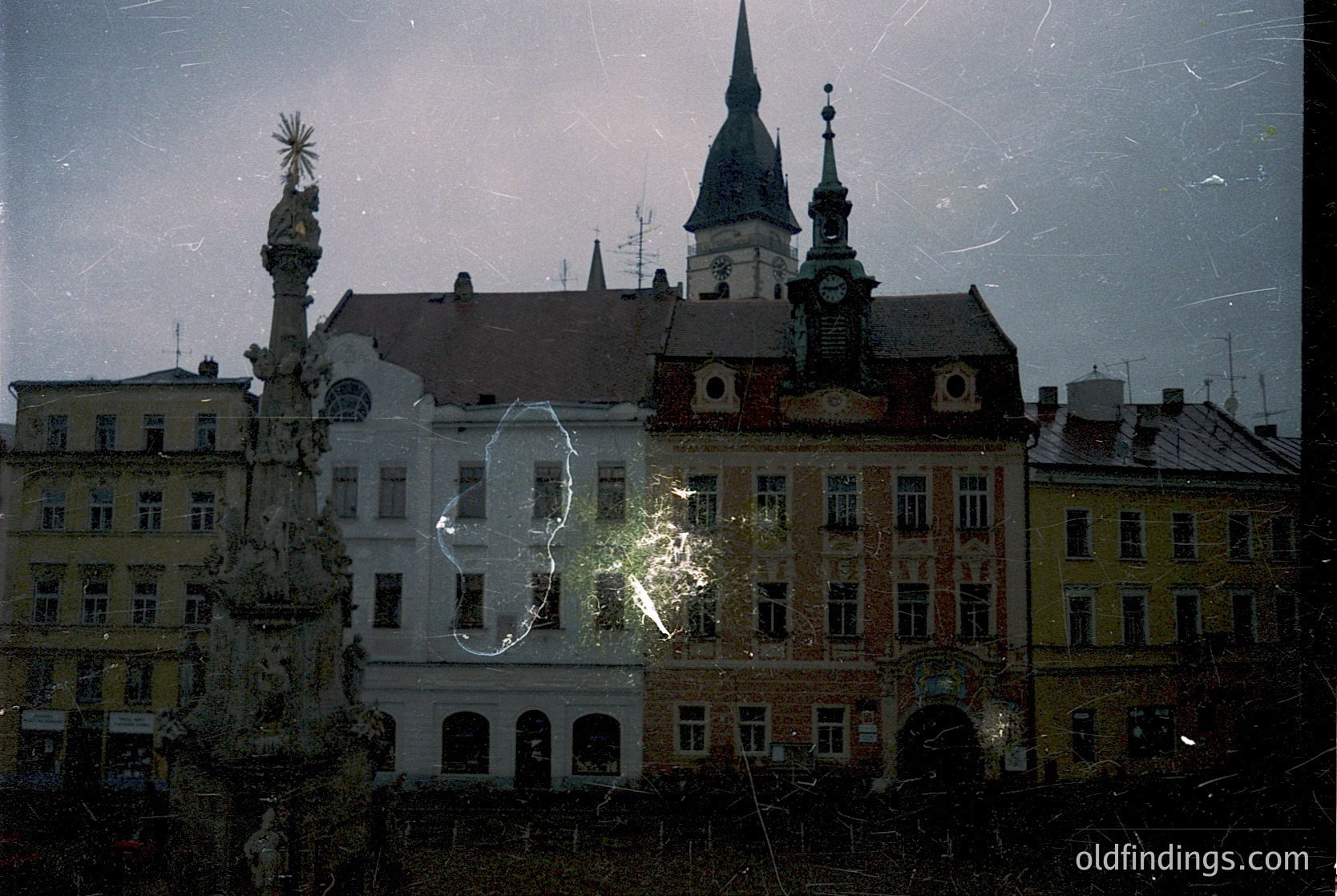 Vintage urban architecture through rain-spattered glass, featuring a Baroque-style fountain with ornate spire and clock tower in background. Likely Eastern European, 1960s-1980s era.