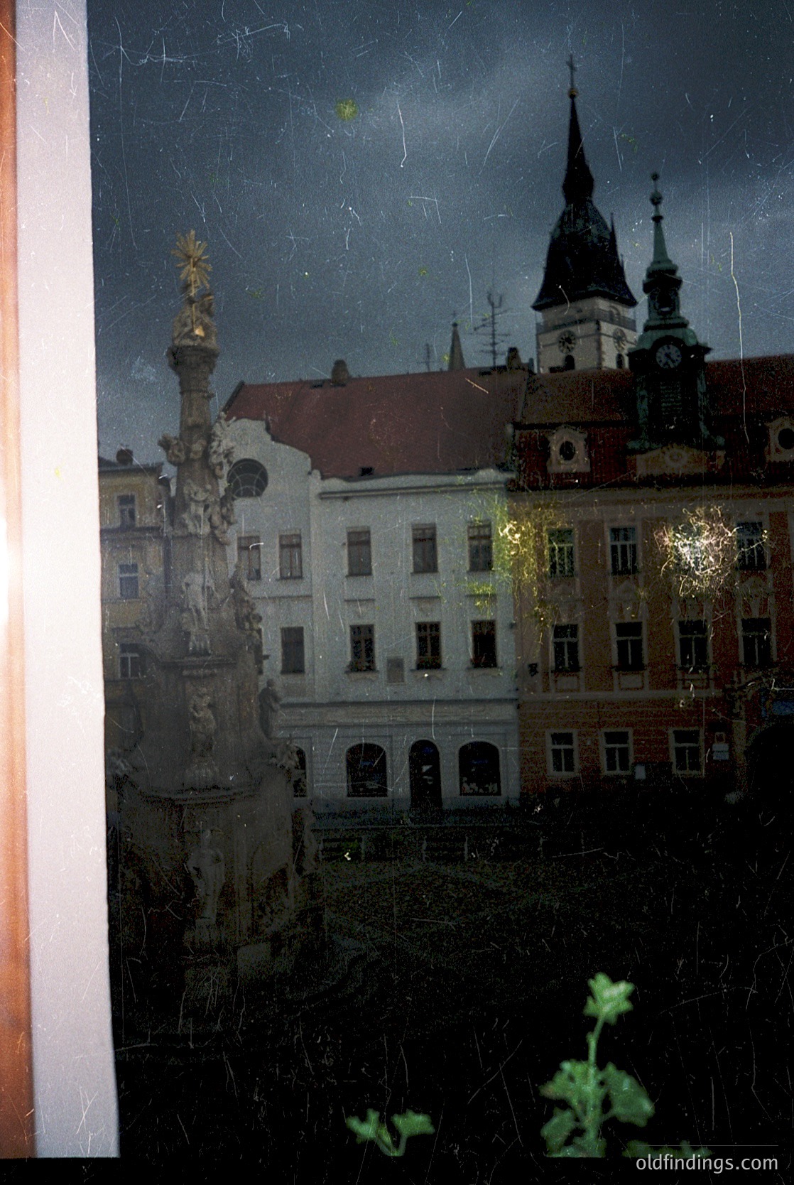 Vintage photograph of a European cityscape viewed through a rain-streaked window. Central column with ornate golden statue atop, flanked by historic buildings with gabled roofs and clock towers. Likely late 19th to early 20th century architecture. Raindrops and condensation blur the scene, adding nostalgic texture.