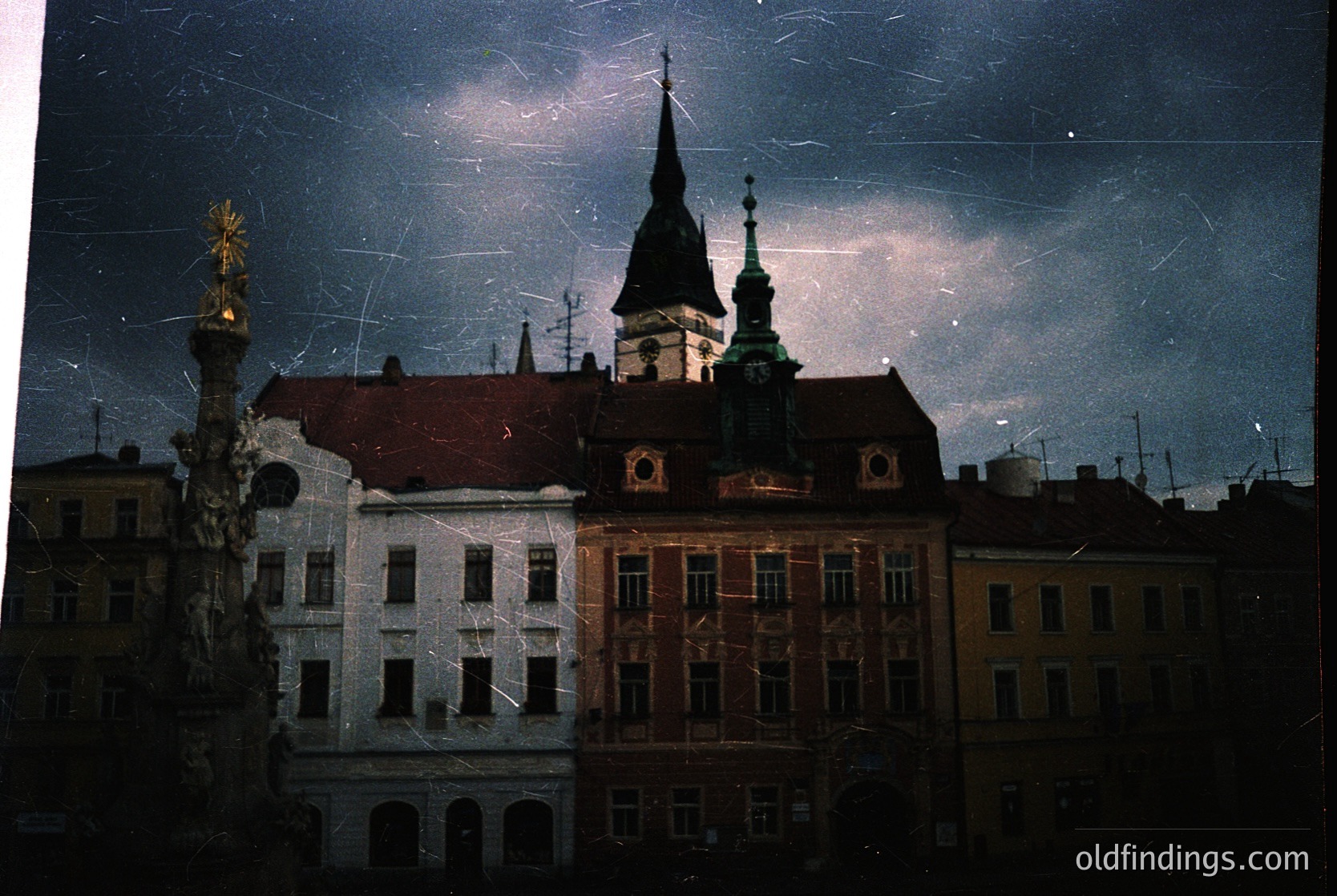 Vintage urban night scene featuring a historic European plaza with a prominent church steeple and ornate column. Architectural details include Baroque-style facades and a dark, moody sky with visible light trails. Likely 20th-century urban photography.