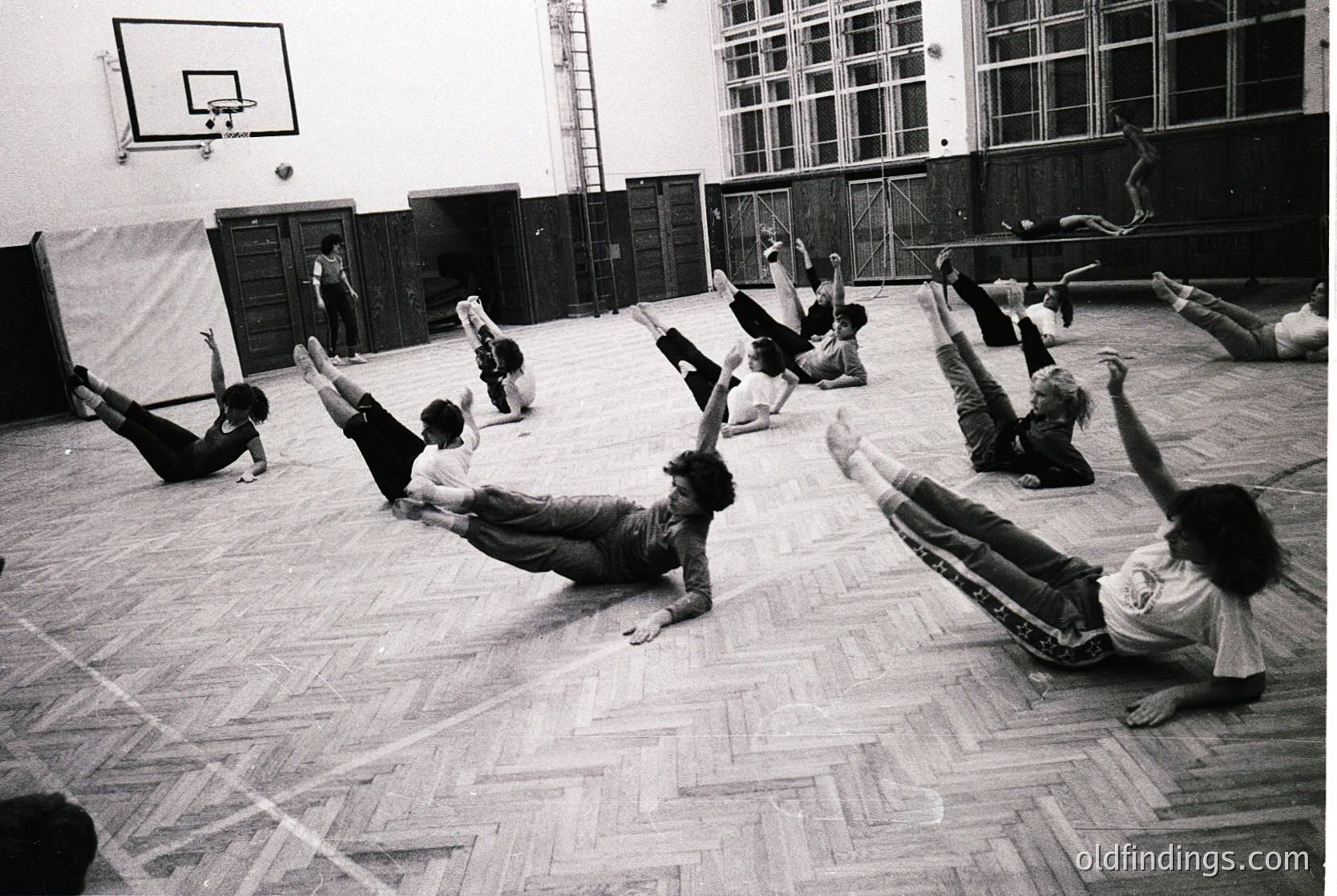 Group fitness routine in a gymnasium, likely 1960s–1980s. Participants perform floor exercises in unison, with one instructor visible at the back. Wooden flooring and industrial-style lockers suggest institutional use. Black-and-white aesthetic highlights dynamic movement and mid-century design.