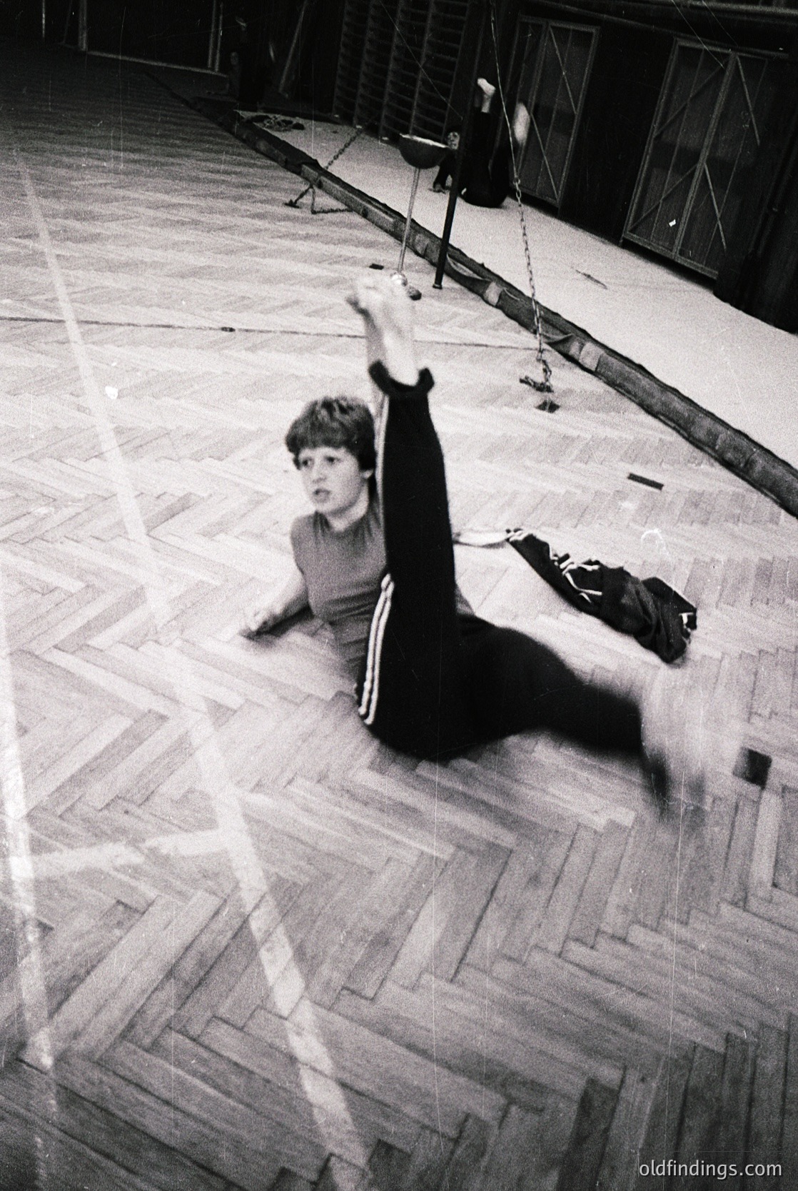 Young gymnast mid-dynamic swing on uneven bars in a vintage indoor gym, 1960s–1970s. Wooden parquet floor, high ceilings, and industrial lighting highlight the era’s sports infrastructure.