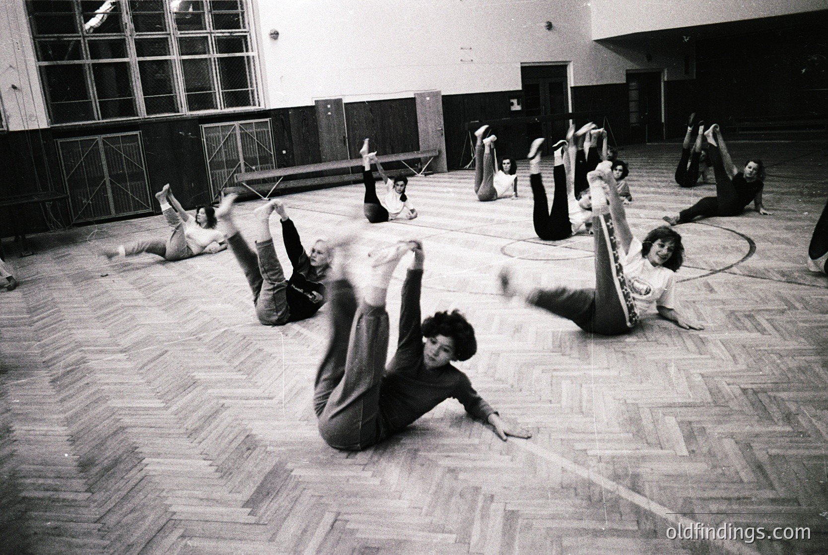 Group gymnastics session in a large indoor hall, likely a school or sports facility. Participants perform floor exercises in mid-motion, 1960s–1970s style. Wooden floor and high windows suggest institutional setting. Dynamic poses capture fluidity of movement.