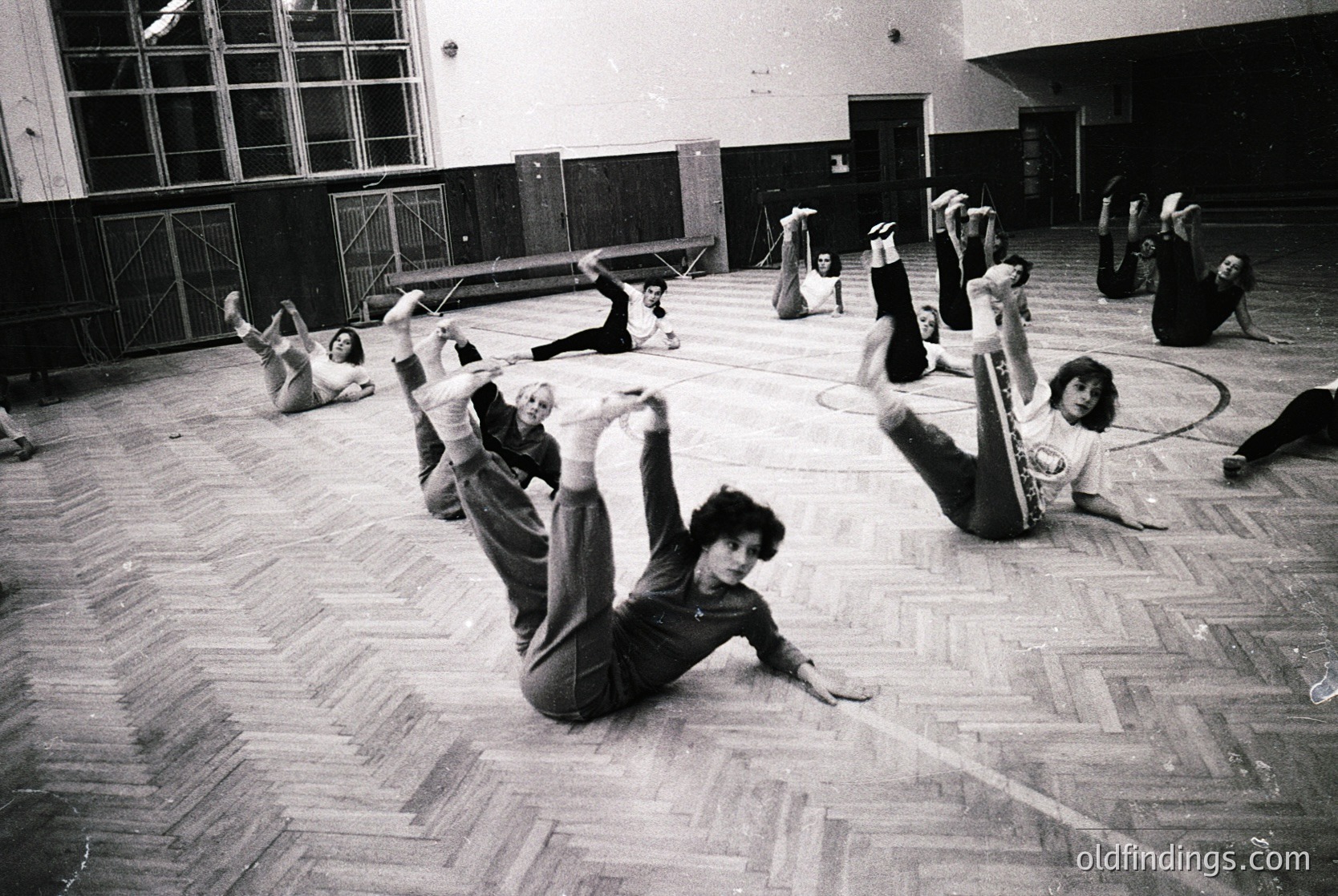Group fitness class in a gymnasium, likely 1970s–1980s. Participants perform floor exercises in a spacious, wood-floored hall with high windows and a stage backdrop. Casual athletic wear and dynamic poses suggest a structured but informal workout session.