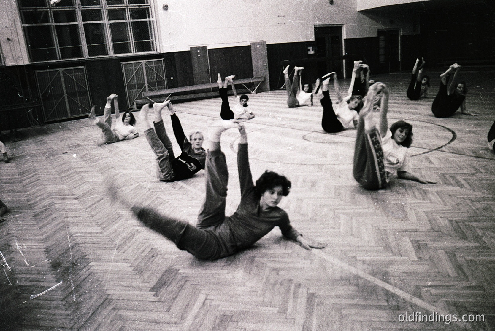 Group exercise class in a large indoor gymnasium, likely 1970s–1980s. Participants perform floor stretches in coordinated poses, emphasizing flexibility. Wooden flooring and high windows suggest institutional or community use.