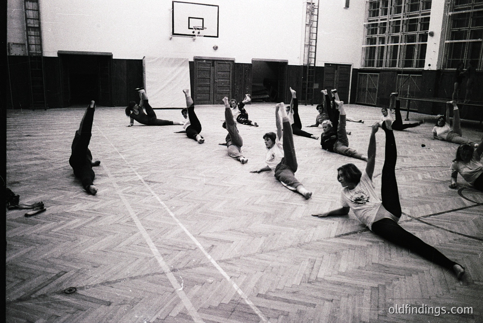 Group fitness class in a gymnasium, likely 1970s–1980s. Participants perform floor exercises in a large, wood-floored hall with a basketball hoop and wooden bleachers. Minimalist, functionalist architecture suggests institutional use.