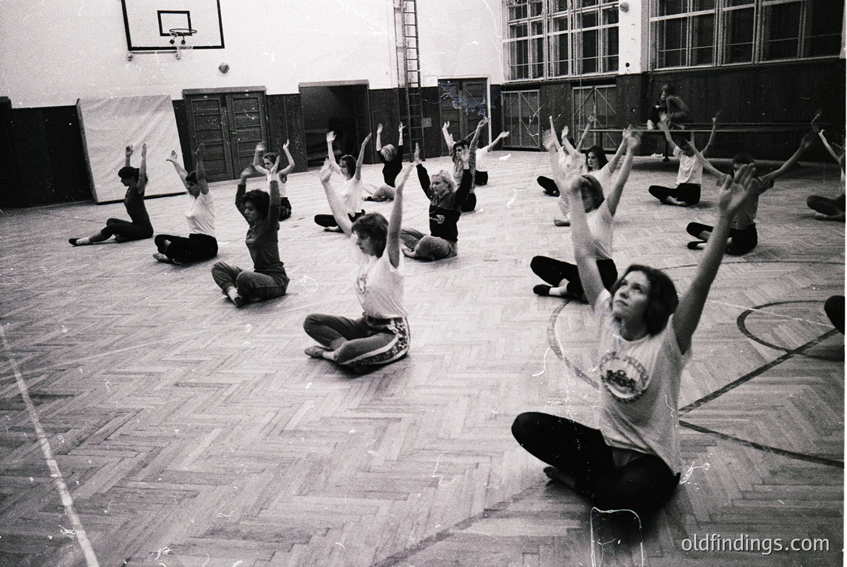 Group fitness class in a gymnasium, likely 1970s–1980s. Participants in coordinated athletic wear perform arm-raising exercises on wooden flooring. Lockers and chalkboards in background suggest institutional use.