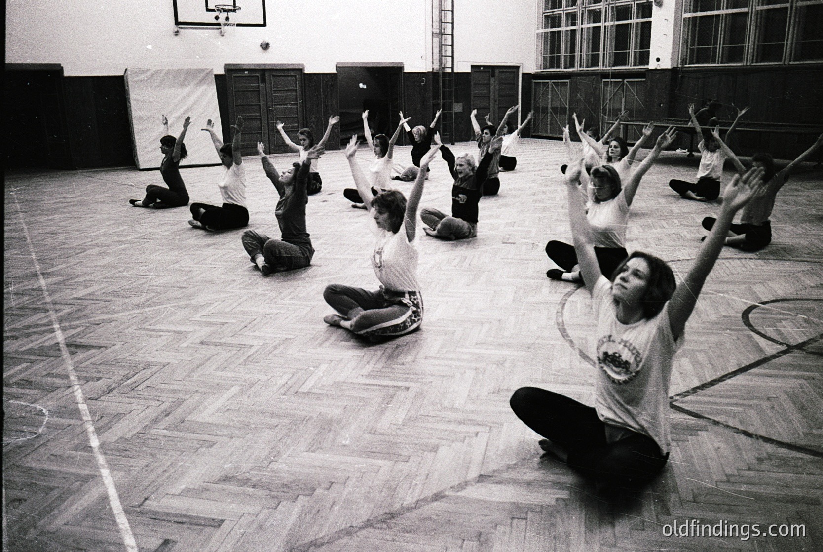 Group exercise class in a gymnasium, mid-20th century. Participants in coordinated athletic wear perform synchronized arm raises. Wooden flooring and vintage lockers in background suggest institutional setting, likely a school or community center.