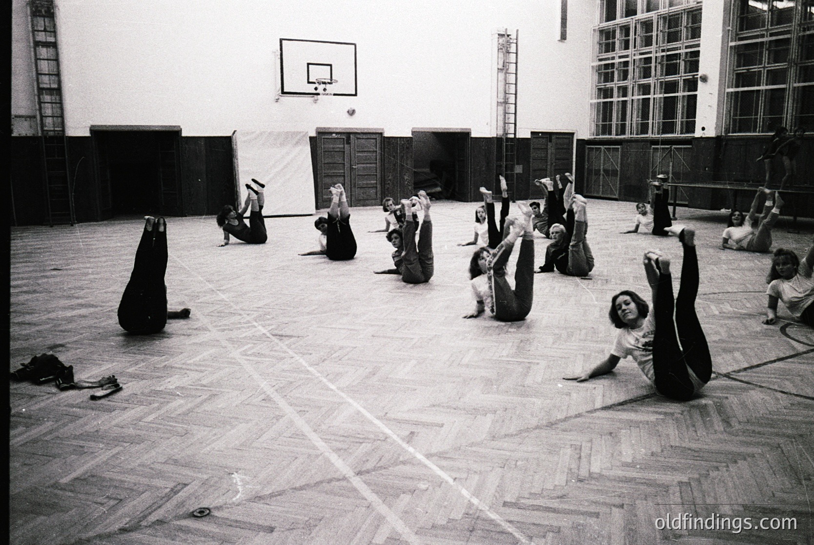 Indoor gymnasium scene with 15+ individuals in dynamic stretching poses, likely a fitness or yoga class. Wooden flooring, basketball hoop, and wooden bleachers in background. Mid-20th century gymnasium design.