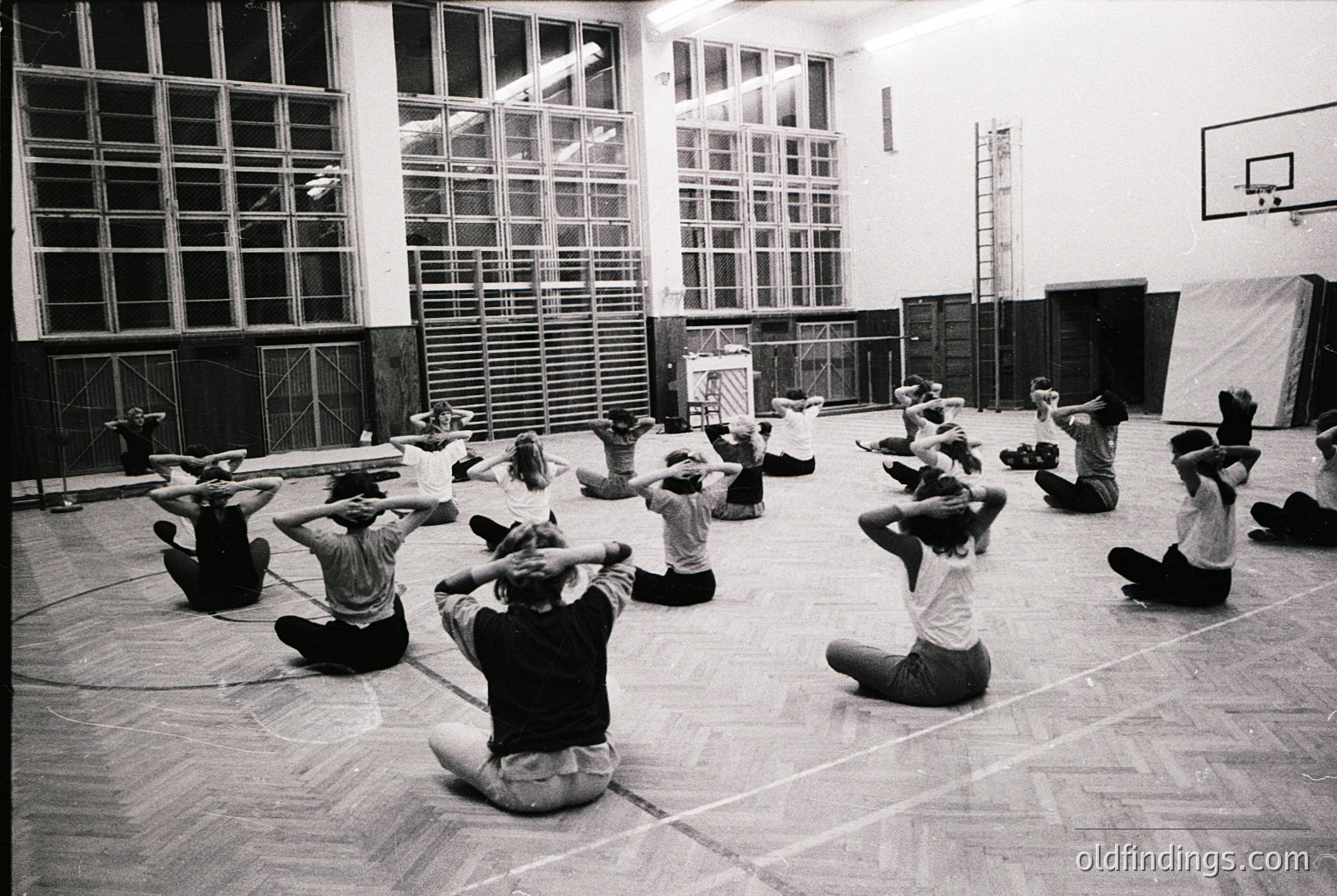 Group fitness session in a large, institutional gymnasium, likely from the 1970s. Participants perform synchronized arm stretches while seated on the floor, under high windows and a basketball hoop. Minimalist decor and wooden flooring suggest a public or educational facility.