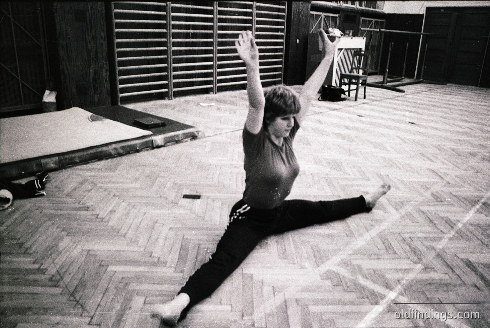 Mid-century dance studio interior with wooden floors, slatted doors, and a trampoline. Female dancer in dynamic split pose, mid-movement, wearing form-fitting leotard and tights. Black-and-white, high-contrast lighting highlights motion and texture. Likely 1960s–1970s, Western Europe/US.