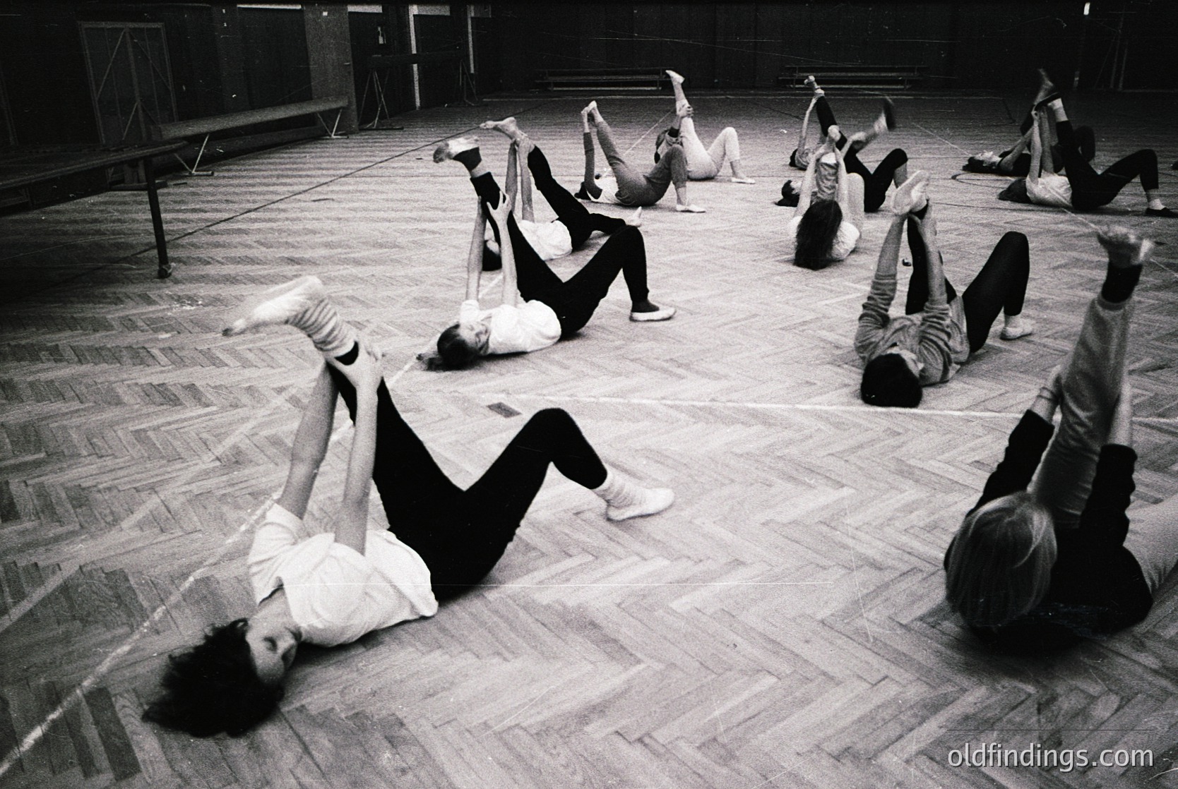 Group performing floor exercises in a spacious, wood-paneled studio—likely a 1960s–1970s dance or gymnastics class. Dynamic poses include splits, backbends, and leg lifts on polished wooden floors. Minimalist attire (leggings, tops) suggests structured movement training.