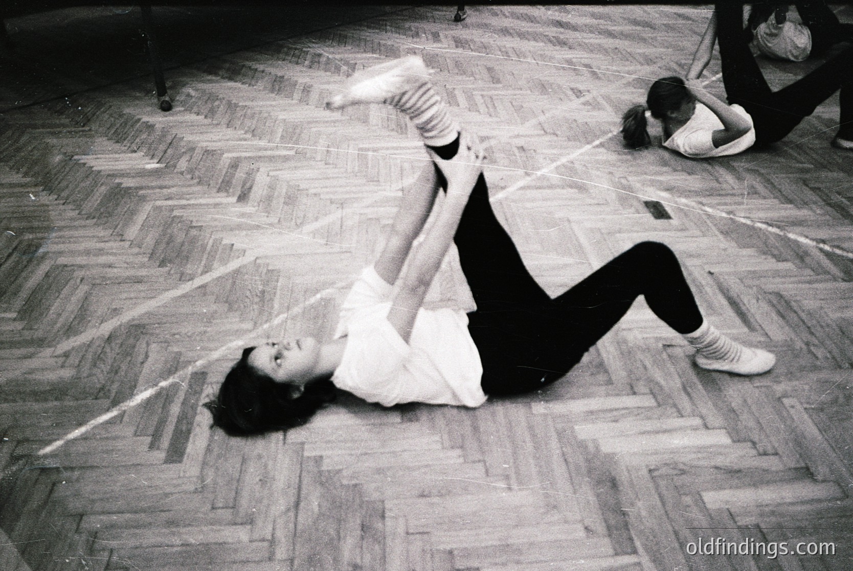 Vintage black-and-white photo of a dynamic floor exercise in a gymnasium, likely mid-20th century. A person performs a split stretch on wooden parquet flooring, wearing a loose blouse and dark pants. Another individual kneels nearby, possibly assisting or observing. Functional, institutional architecture with minimal decor.