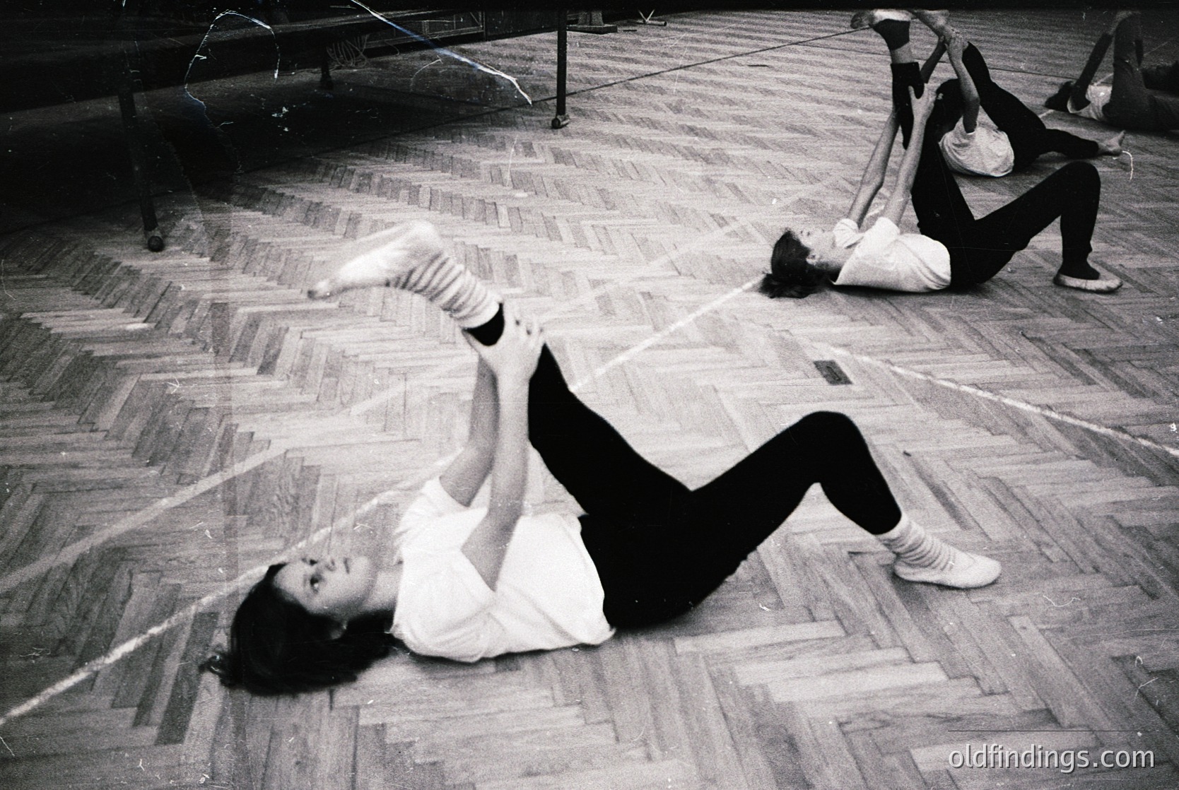 Black-and-white gym scene featuring two individuals performing floor exercises in a wooden-floored hall, likely from the 1960s–1970s. Foreground subject in dynamic split pose; background shows another in seated leg stretch. Industrial pipes and beams suggest institutional or public facility.