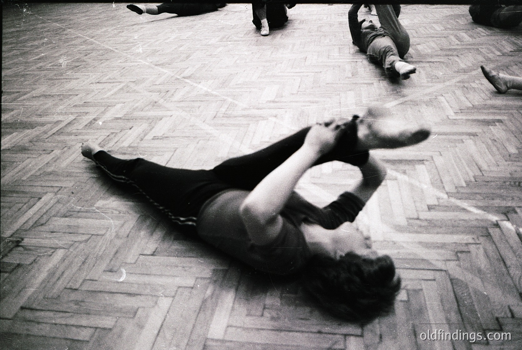 Black-and-white street photography captures a dynamic moment of a person mid-fall on polished wooden floors, likely in a public or commercial space. The individual’s dynamic pose and blurred motion suggest spontaneous movement. Surrounding figures appear relaxed, seated or lying on the floor. Style hints at 1960s–1970s urban culture, possibly European or North American.