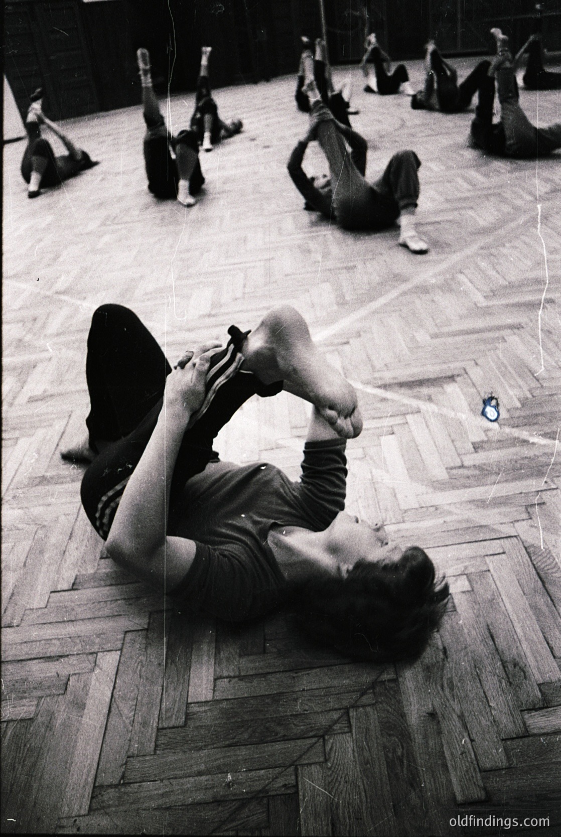 Group fitness class in a gymnasium, 1960s–1970s. Participants perform floor exercises in coordinated poses, emphasizing flexibility and group coordination. Wooden parquet flooring and numbered markers suggest structured routines. Ideal for vintage fitness, physical education, or historical exercise research.