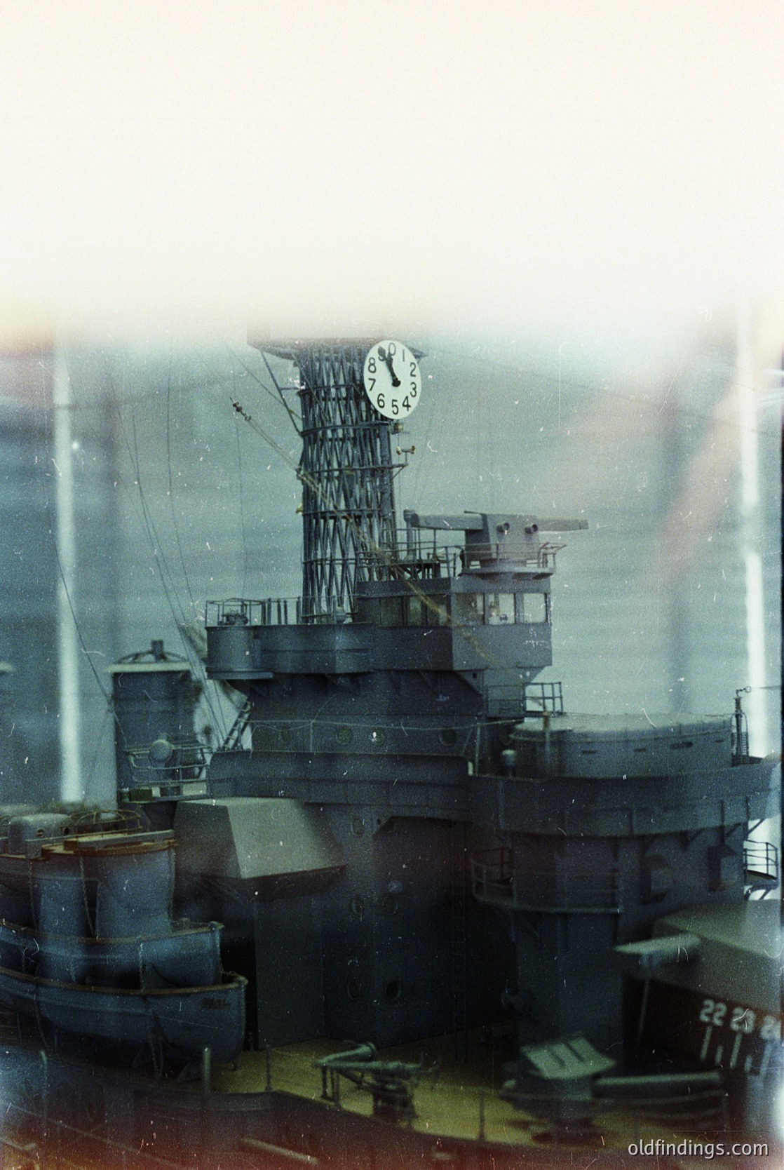 Mid-20th century naval vessel with prominent clock tower and anti-aircraft mounts, likely a WWII-era battleship. Rust and weathering indicate prolonged exposure. Industrial port or naval base setting.