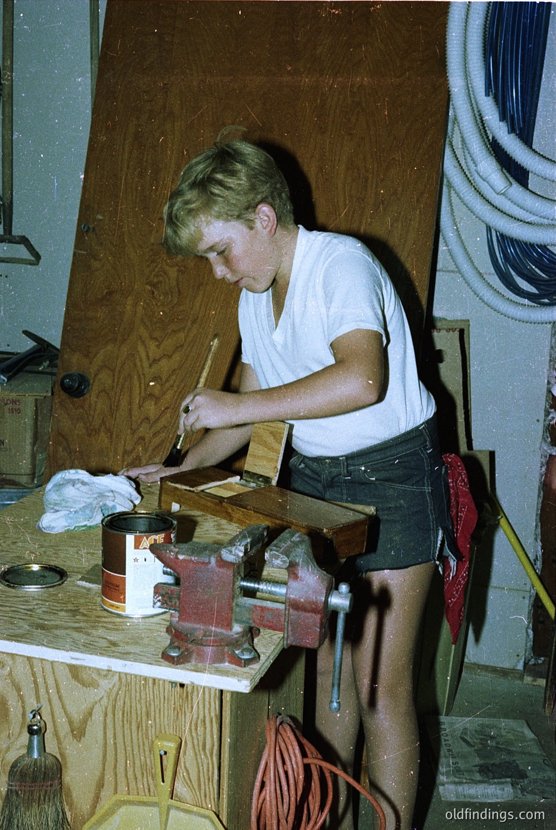 Young man using a hand plane on a wooden workbench in a workshop, likely mid-20th century. Visible tools include a vise, sandpaper, and a red toolbox. Casual attire suggests a DIY or hobbyist setting.