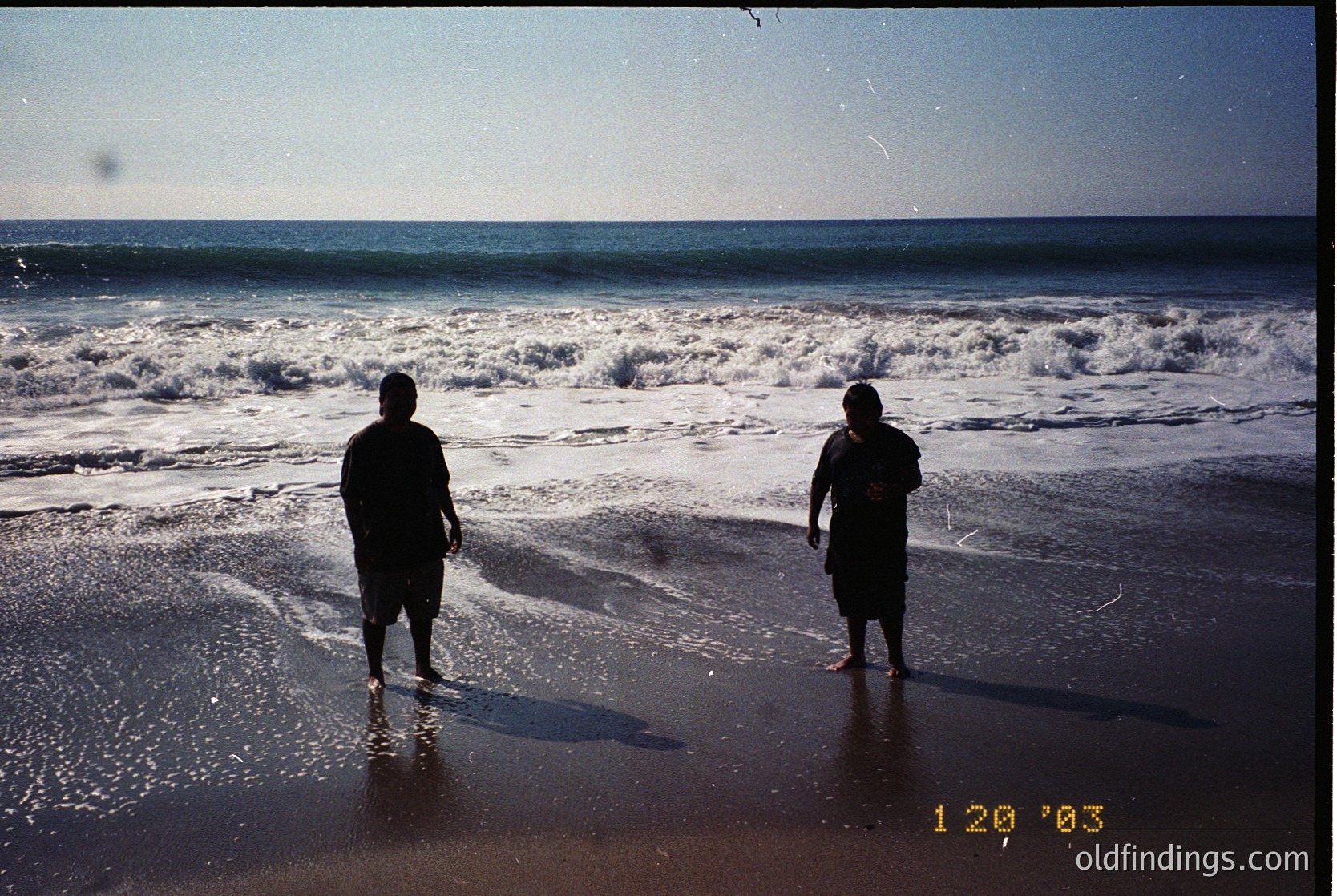 Two figures stand on a wet beach shore, facing crashing waves under a clear sky. Mid-2000s coastal scene with vintage film grain.