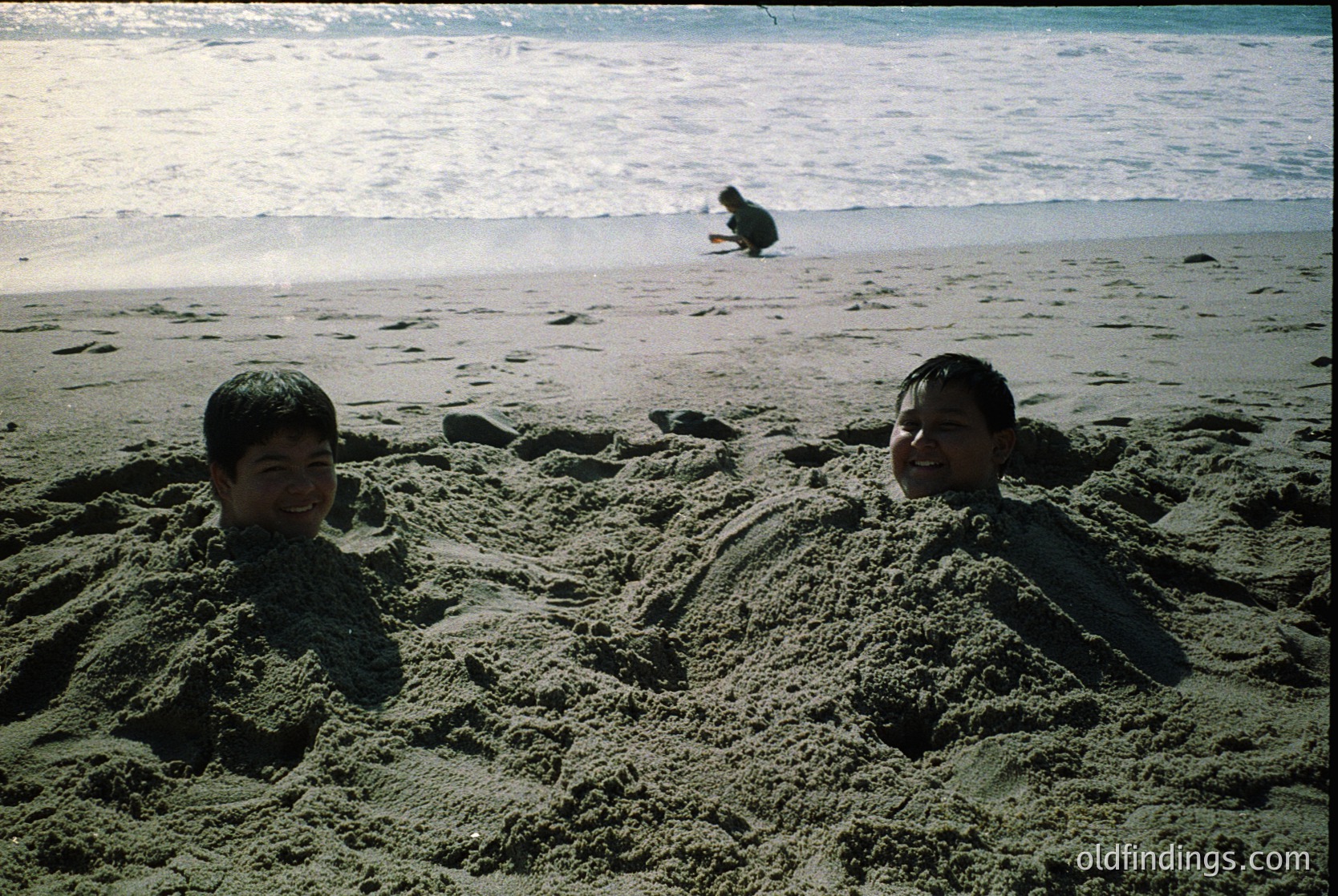 Two children play in a sandcastle on a beach, framed by vintage film grain. Waves crash in the background, and another figure sits farther along the shore. Likely mid-20th century, coastal setting.