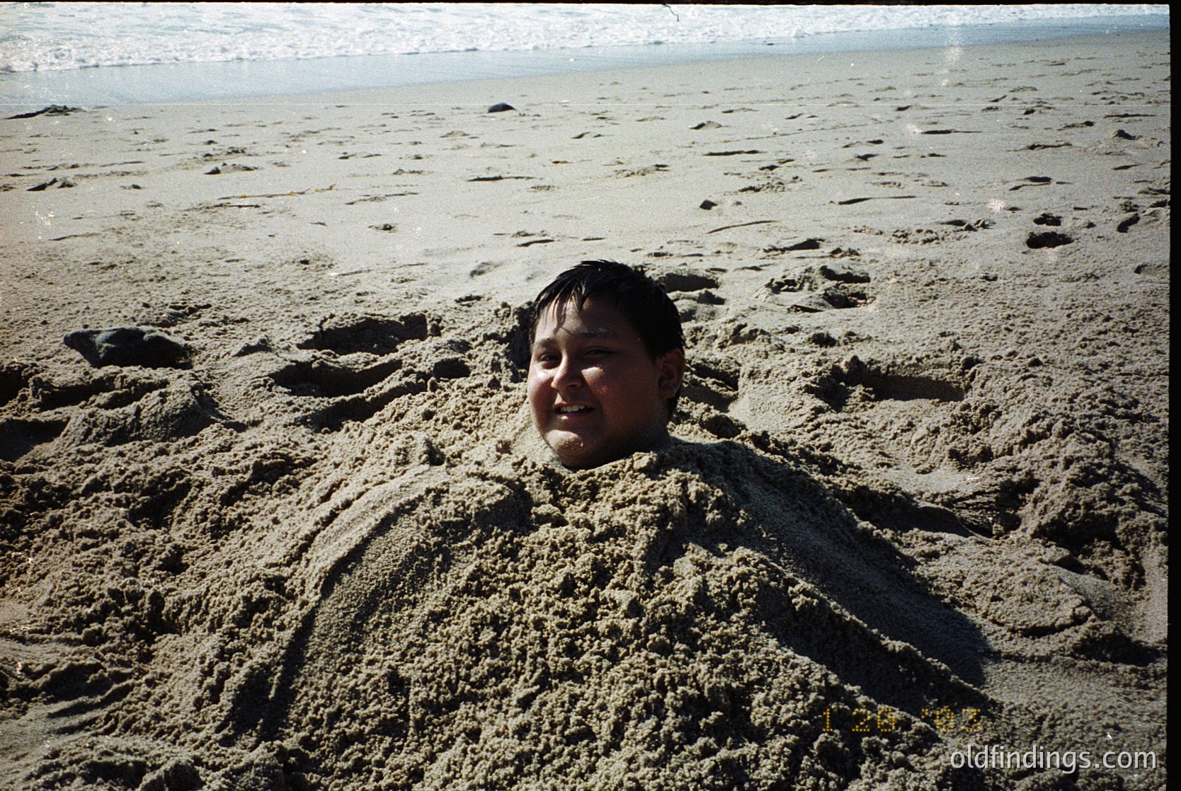 Child partially buried in sand on a wide, empty beach with gentle waves in background. Likely mid-20th century due to film grain and clothing style. Evokes nostalgia and childhood play.