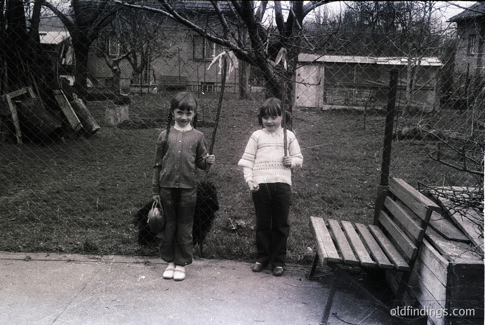Two children pose outdoors in a residential backyard, likely mid-20th century. The girl on the left holds a broom and a bag, while the other carries a knitted sweater. A wooden bench and fenced garden area with a shed are visible. Urban residential setting, early school-age attire.