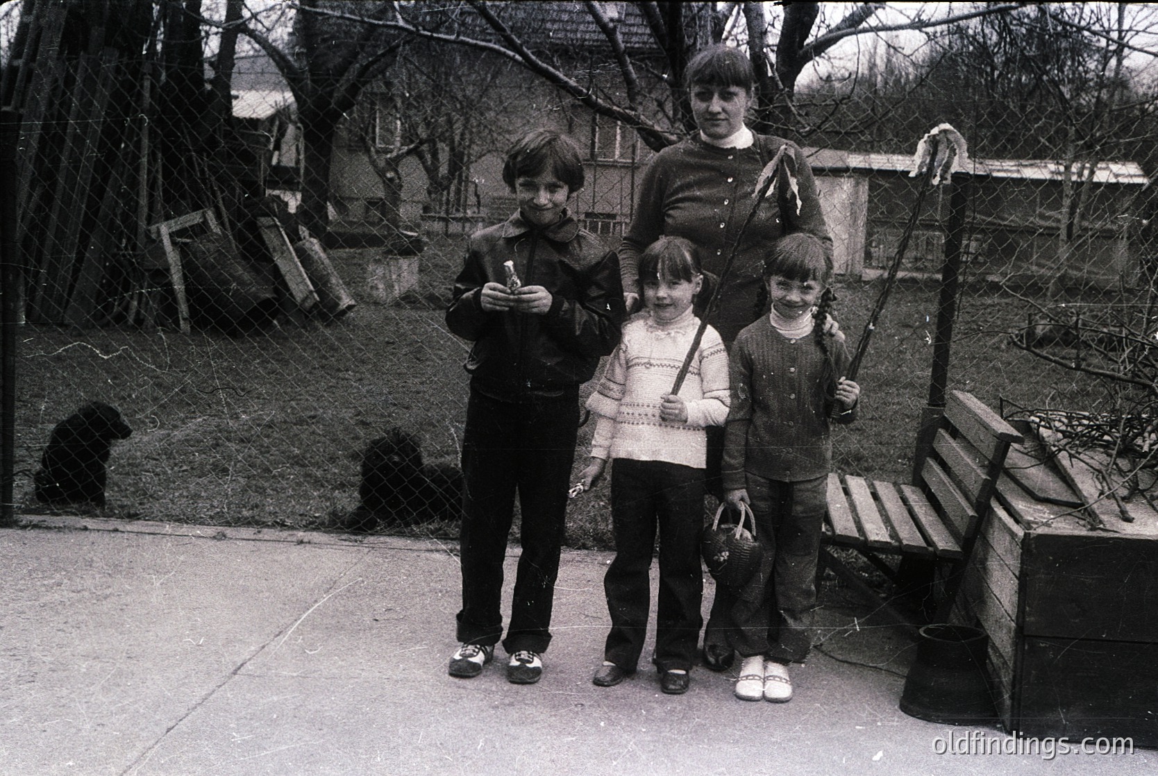 Family portrait in a residential courtyard, likely 1970s–1980s. Four individuals pose outdoors near a wooden bench and rustic fence. The adult wears a long coat and holds a cane; children in layered sweaters and trousers. Leaves suggest autumn or early winter. Urban residential setting.