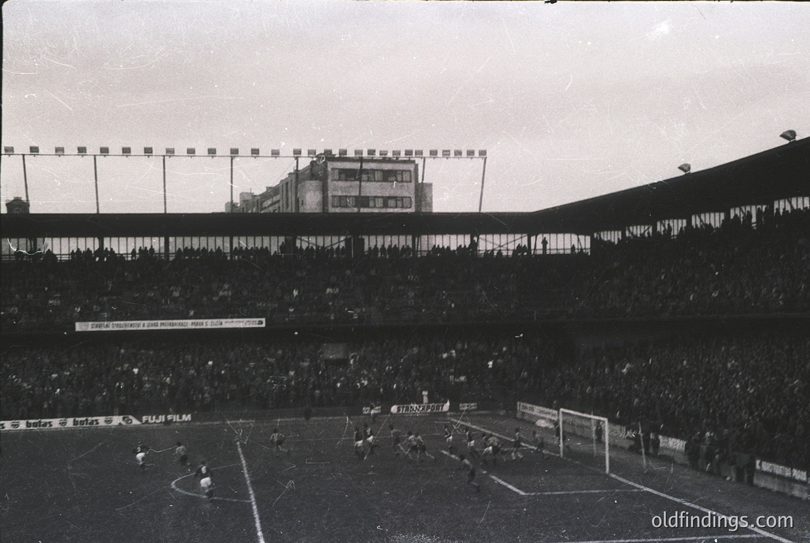 Vintage black-and-white stadium shot showing a packed arena with tiered seating and a sparse field. Visible signage includes "Fuji Film" and Cyrillic text, suggesting Eastern European setting. Crowd density and stadium design hint at mid-20th century sports culture.