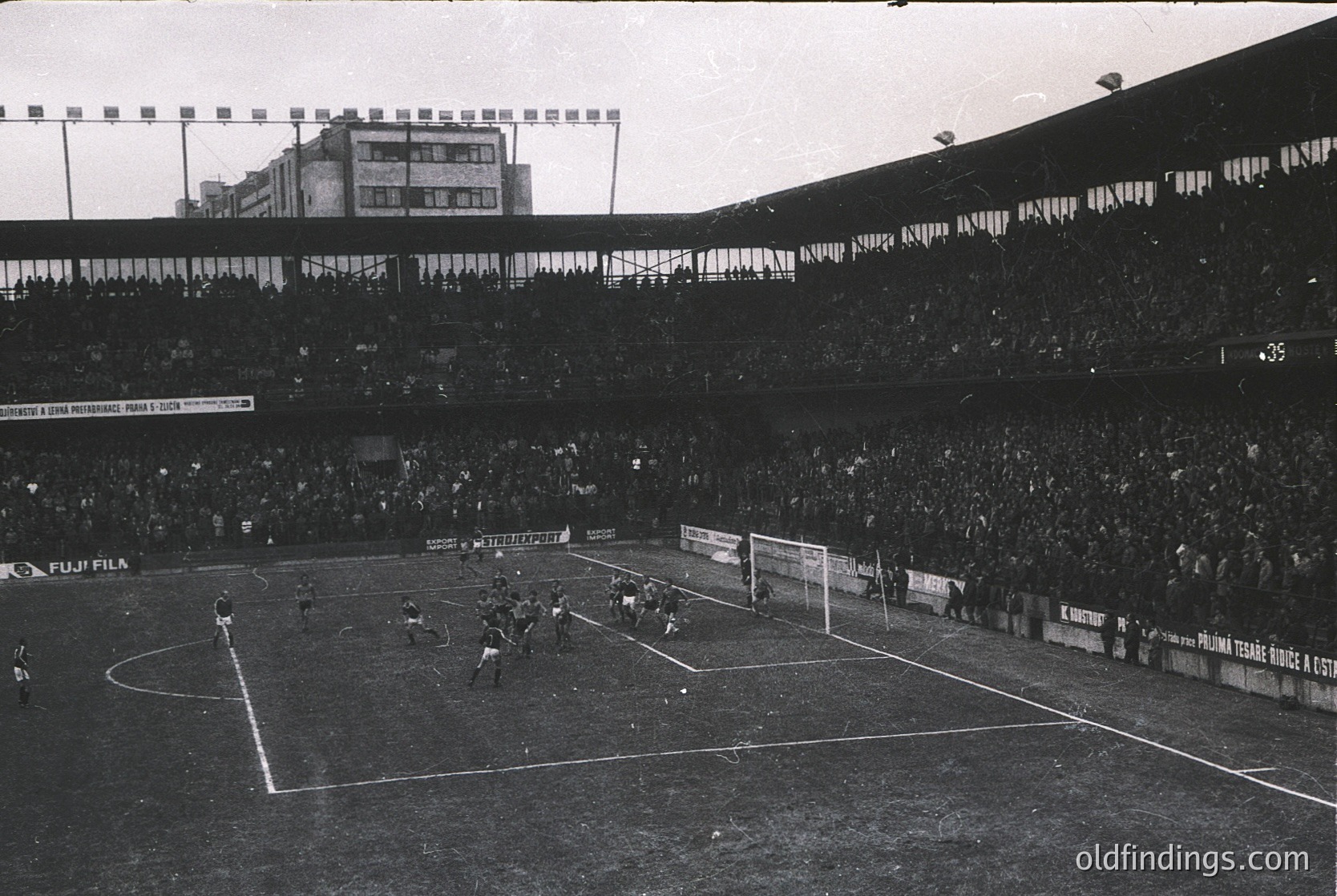 Black-and-white stadium shot capturing a 1970s European football match. Crowded stands with dense seating, visible advertisements like *Puma* and *Kronenbourg*, and a packed pitch with players in action. Urban backdrop of multi-story buildings.