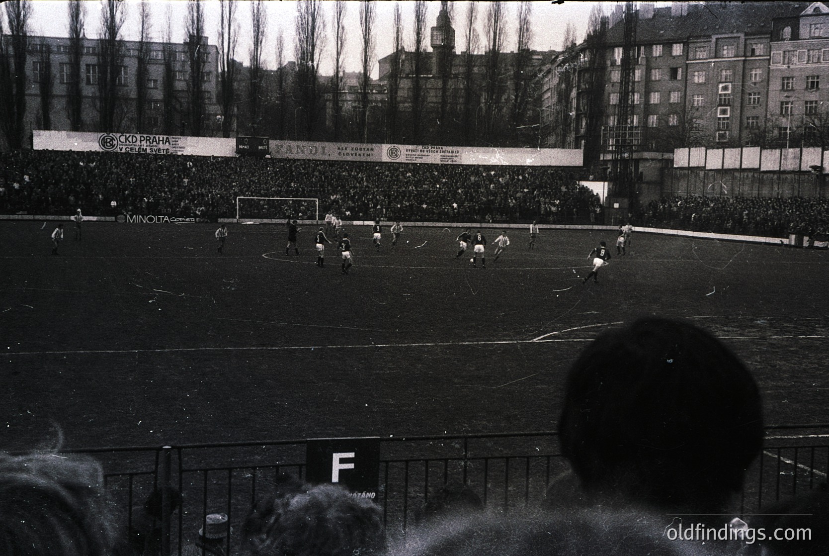 Black-and-white shot of a packed stadium during a 1960s–70s soccer match, likely in Czechoslovakia ( ). Crowds fill stands behind barricades, with visible sponsors like "Minolta" and "Spartak" on banners. Urban architecture with multi-story residential blocks frames the scene. Crowd perspective from lower tier reveals dense, enthusiastic attendance.