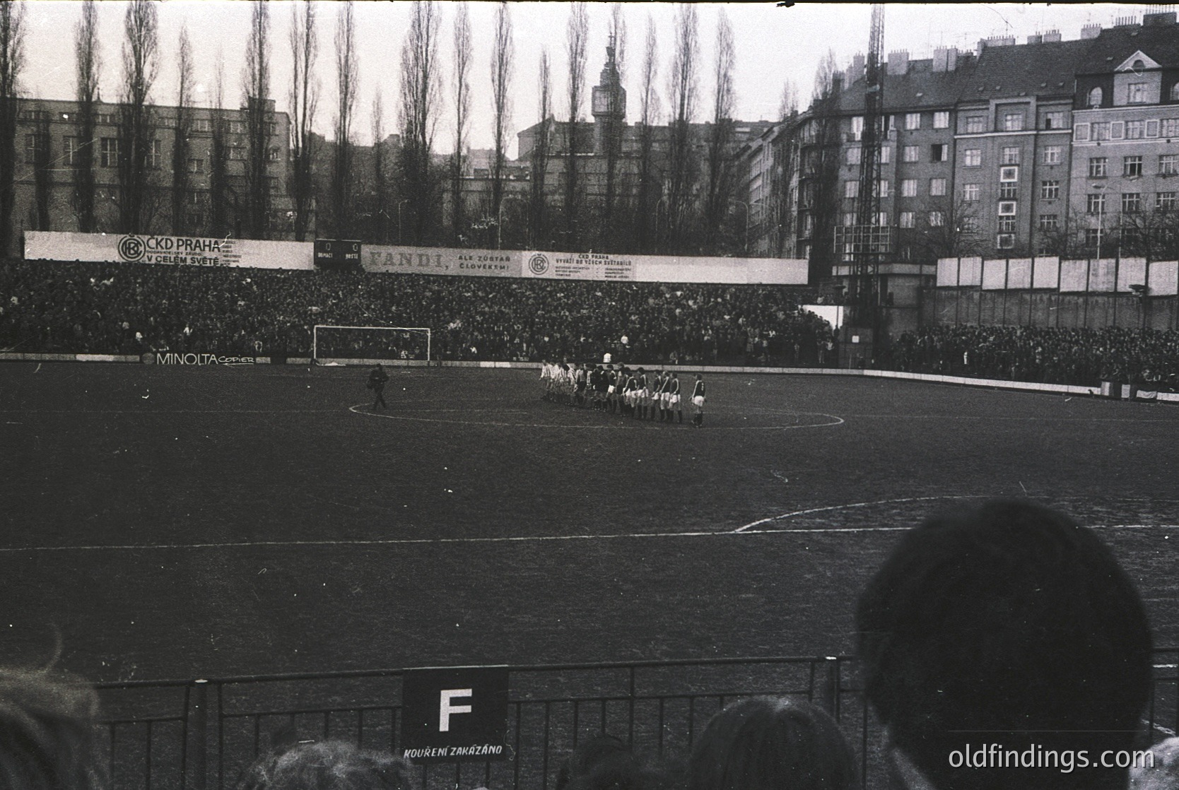 Black-and-white photo of a packed stadium during a 1960s football match. Crowds line the perimeter, with visible banners advertising brands like *Kupfer* and *Andi*. Players in white uniforms play on a grass field, framed by urban architecture and leafless trees. Crowd density suggests high attendance.