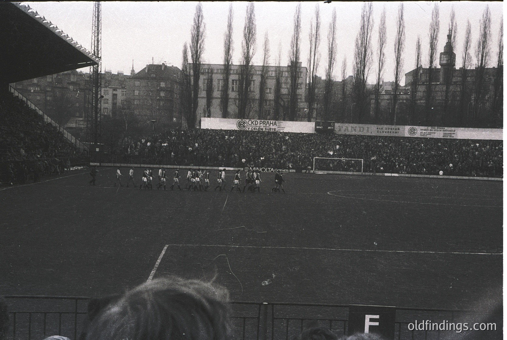 Black-and-white shot of a packed European stadium, likely –70s, featuring a packed crowd and a match in progress. Banner ads for brands like "Tandi" and "Cordura" suggest mid-century commercial sponsorship. Urban backdrop with dense residential buildings and a visible church spire. Crowd density and stadium design reflect era’s football culture.