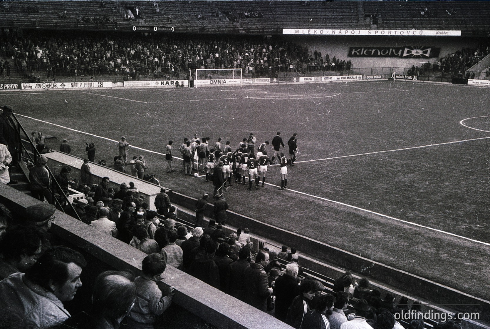 Black-and-white shot of a mid-20th century football match at a packed stadium, likely Eastern Bloc era. Players in white kits line up for kickoff; spectators fill stands, some holding flags. Advertisements for brands like *Renault* and *Omnia* visible. Crowd density suggests high attendance.