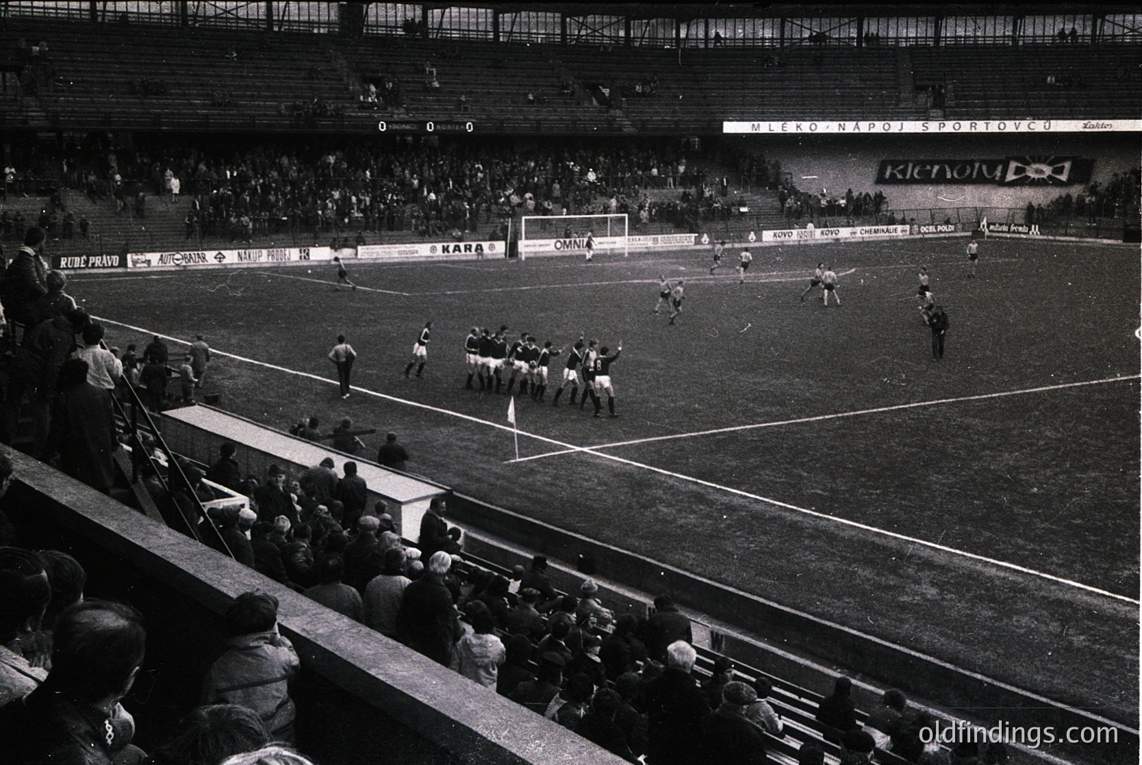 Black-and-white shot of a packed stadium during a mid-20th-century football match, likely Eastern Bloc era. Players in white and dark uniforms clash near the center circle, while spectators fill tiered stands. Advertising banners (e.g., "Kara") and Soviet-style signage ("Унион Спорт") suggest a Bulgarian venue. Crowded atmosphere reflects mid-1960s–70s European football culture.