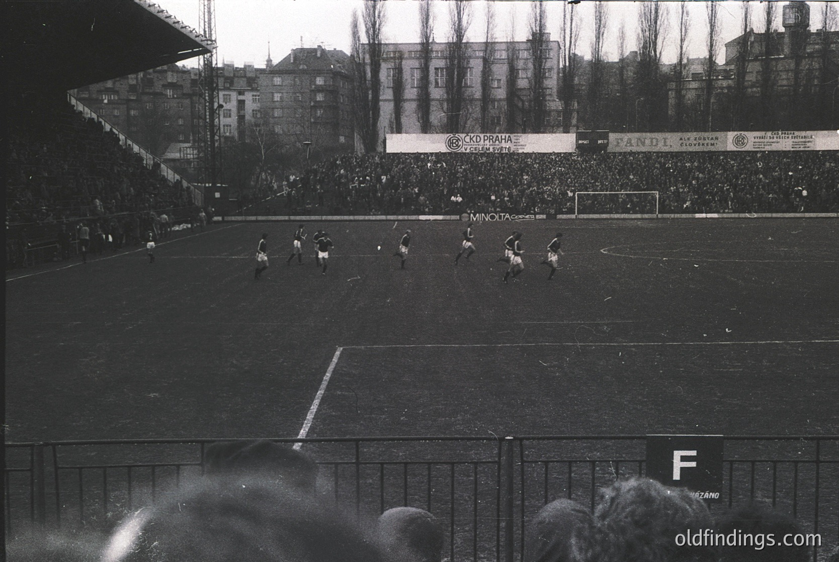 Vintage black-and-white soccer match in a packed stadium, featuring mid-action players on a grass field. Advertising banners (Minota, Fandi) and dense crowd in stands suggest 1960s–1970s European league. Urban backdrop of multi-story buildings. Ideal for sports history, retro design, or cultural research.