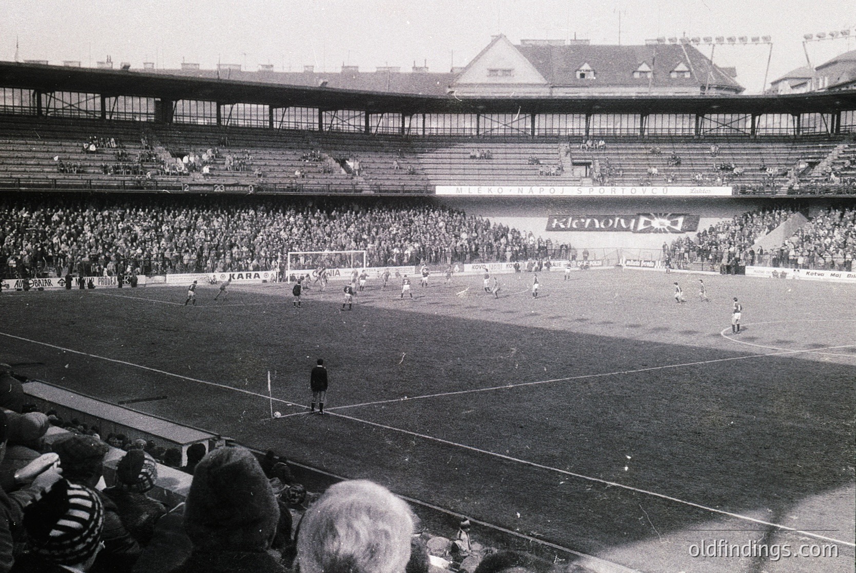 Vintage black-and-white shot of a packed stadium during a football match, likely 1960s–1970s. Multi-tiered stands filled with spectators, central pitch with players in action, and visible "Kendall" branding on a board. Architectural details include curved roofing and tiered seating.