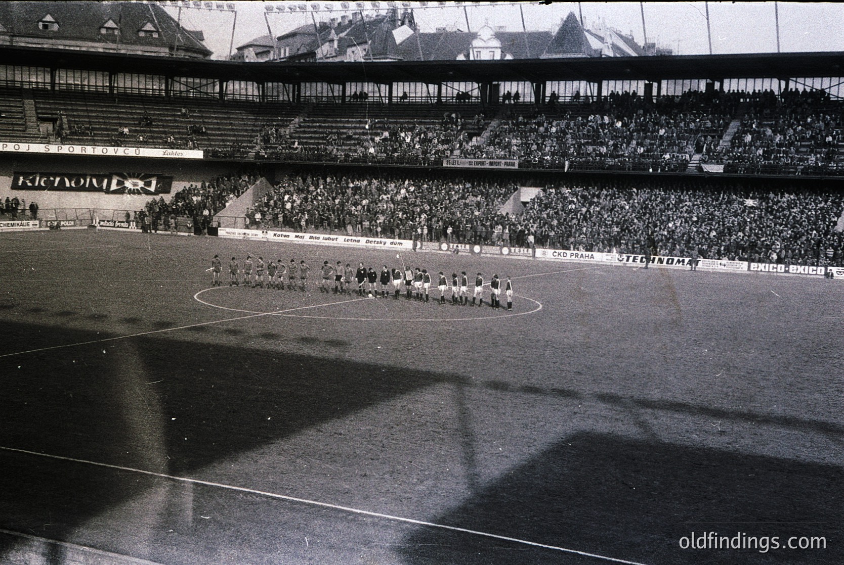 Black-and-white stadium shot showing a pre-match ceremony with teams lined up on a wet pitch. Multi-tiered seating filled with spectators, visible branding like "Steinlager" and "Exxon." Architectural details include tiered concrete stands and a distant rooftop. Likely European, mid-20th century (1960s-1970s).