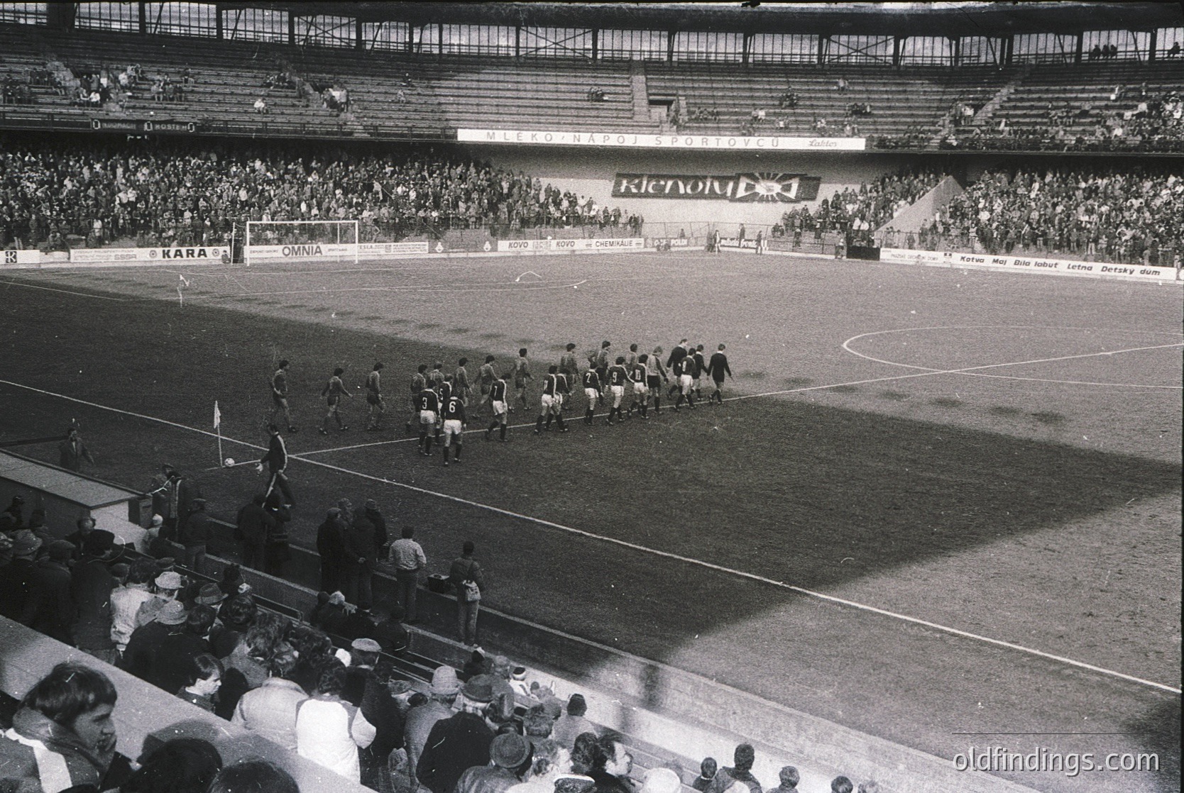 Black-and-white stadium shot showing pre-match team lineups on a grass pitch, flanked by tiered seating filled with spectators. Advertisements for brands like "Kara" and "Iskra" visible on boards. Crowd appears dense, indicating a major event. Mid-20th century European football atmosphere.