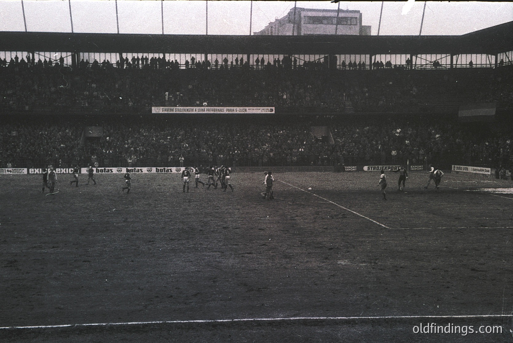 Black-and-white soccer match in a packed stadium, likely 1960s–1970s. Players in white and dark jerseys compete on a muddy field under overcast skies. Advertisements for "BX1 FABXIC" and "Botas" line the perimeter. Dense crowd fills tiered stands, suggesting high attendance. Urban backdrop hints at European setting.