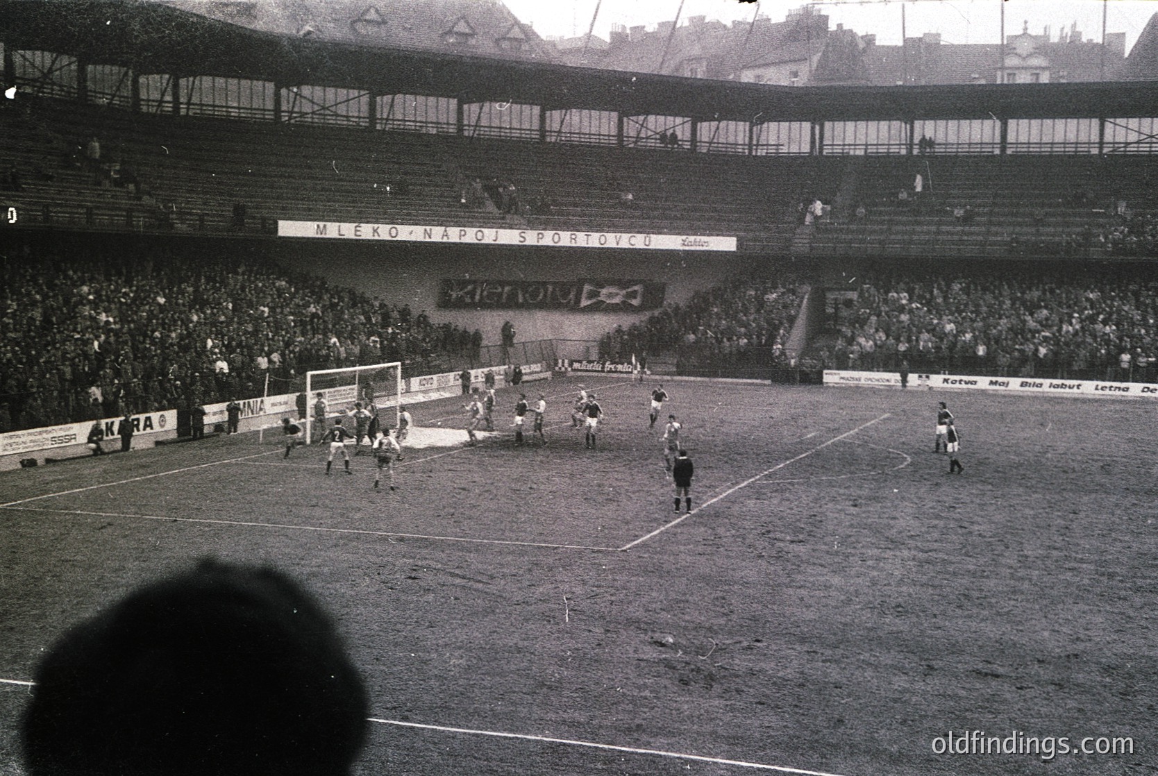 Black-and-white soccer match at a packed stadium, featuring a mid-action play near the goal. Crowds fill tiered stands with banners reading "MĚRO NÁROD SPORTOVCŮ" (Czech: "City of Sports Fans"). Referees, players, and spectators create dynamic tension. Architectural details include high seating tiers and advertising boards. Likely 1960s–1970s, Eastern Bloc region.