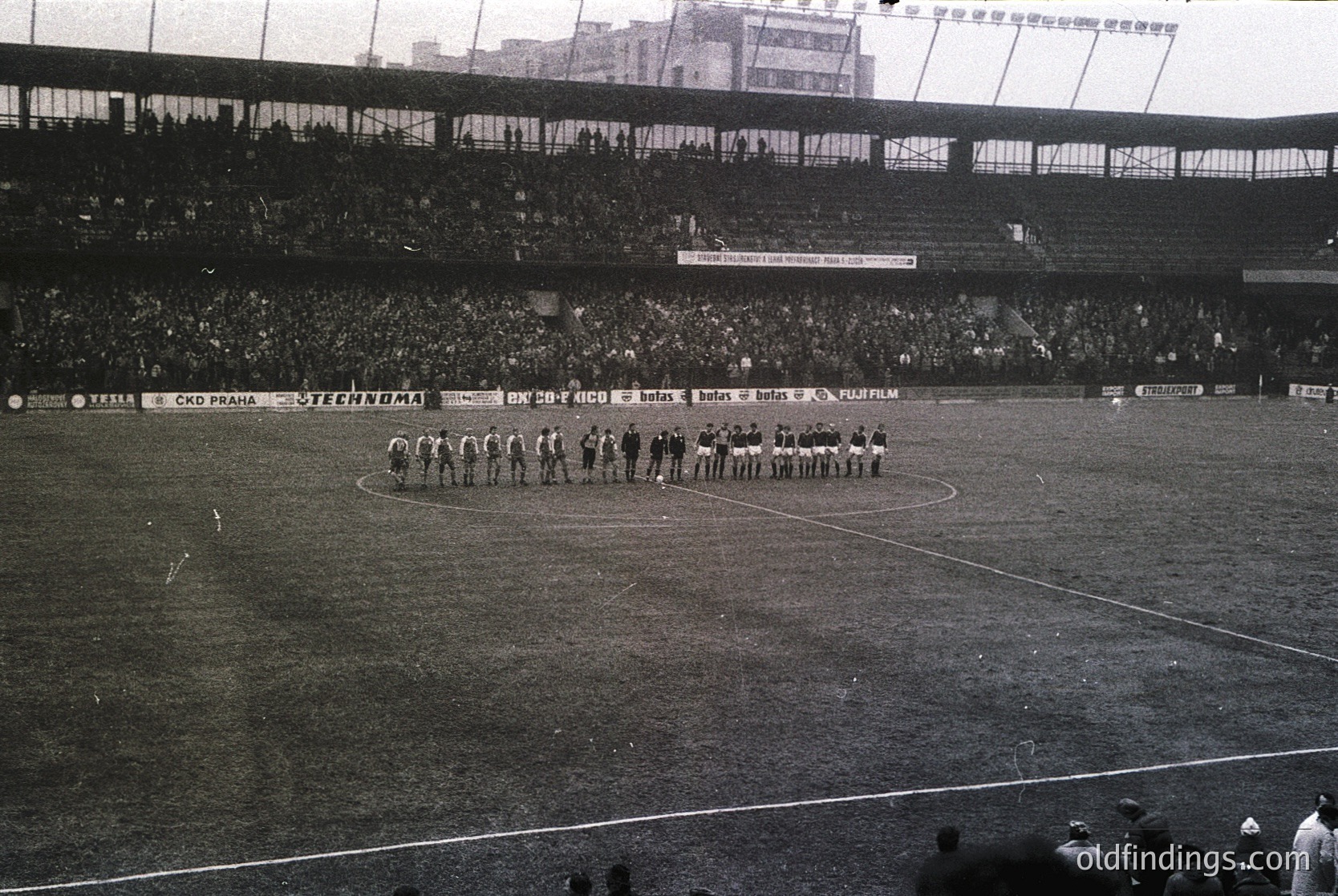 Black-and-white stadium photo showing two football teams in pre-match formation, 1960s-70s. Teams stand on a grass pitch with banners advertising brands like *Steinhof* and *Fulfilm*. Crowd fills tiered seating, suggesting a major event. Architectural details include concrete stands and floodlights.