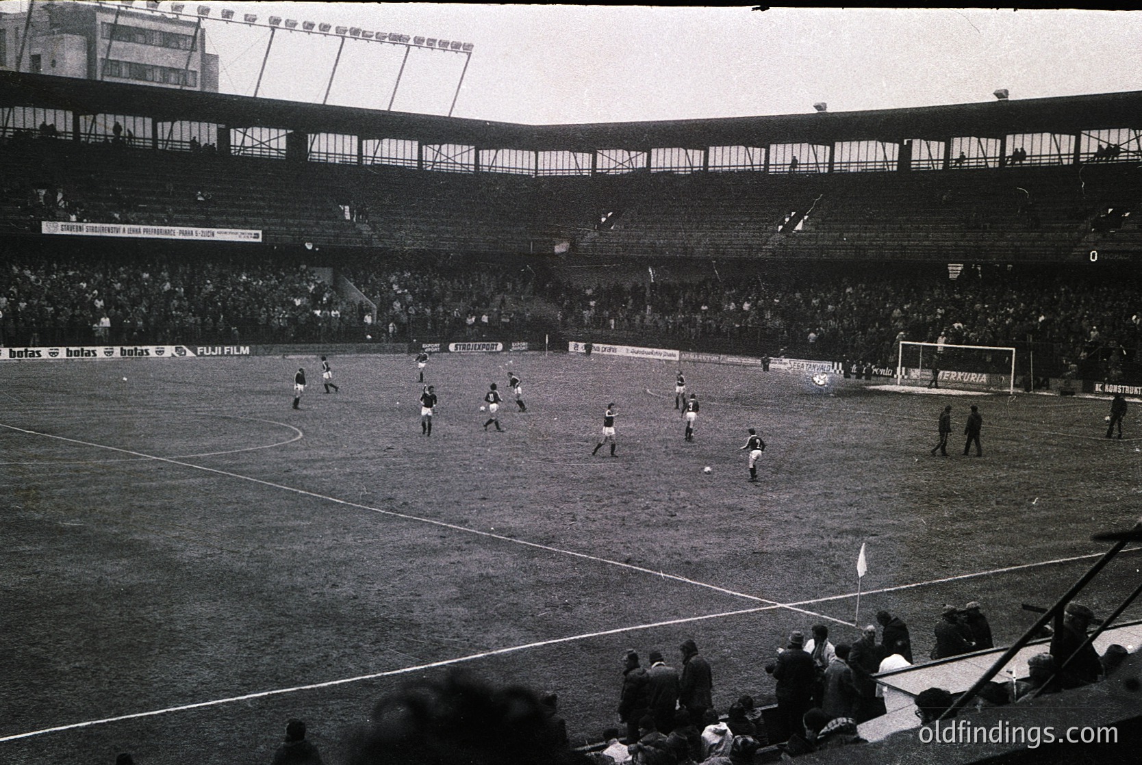 Black-and-white stadium shot featuring a mid-20th century football match. Players in dark uniforms chase the ball on a grass pitch under floodlights, with sparse spectators in stands. Advertising banners (Fujifilm, Betasom) and stadium signage visible. Likely European, 1960s–1970s era.